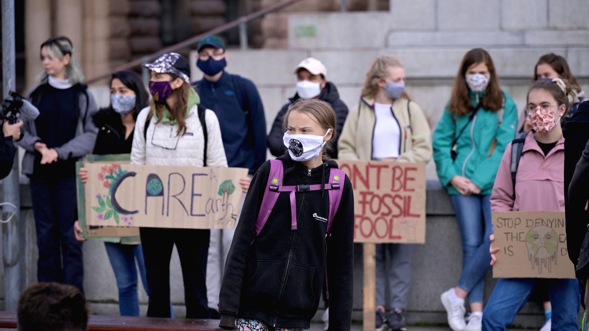 Swedish climate change activist Greta Thunberg is shown standing among a group of activists and wearing a purple backpack and a face mask.