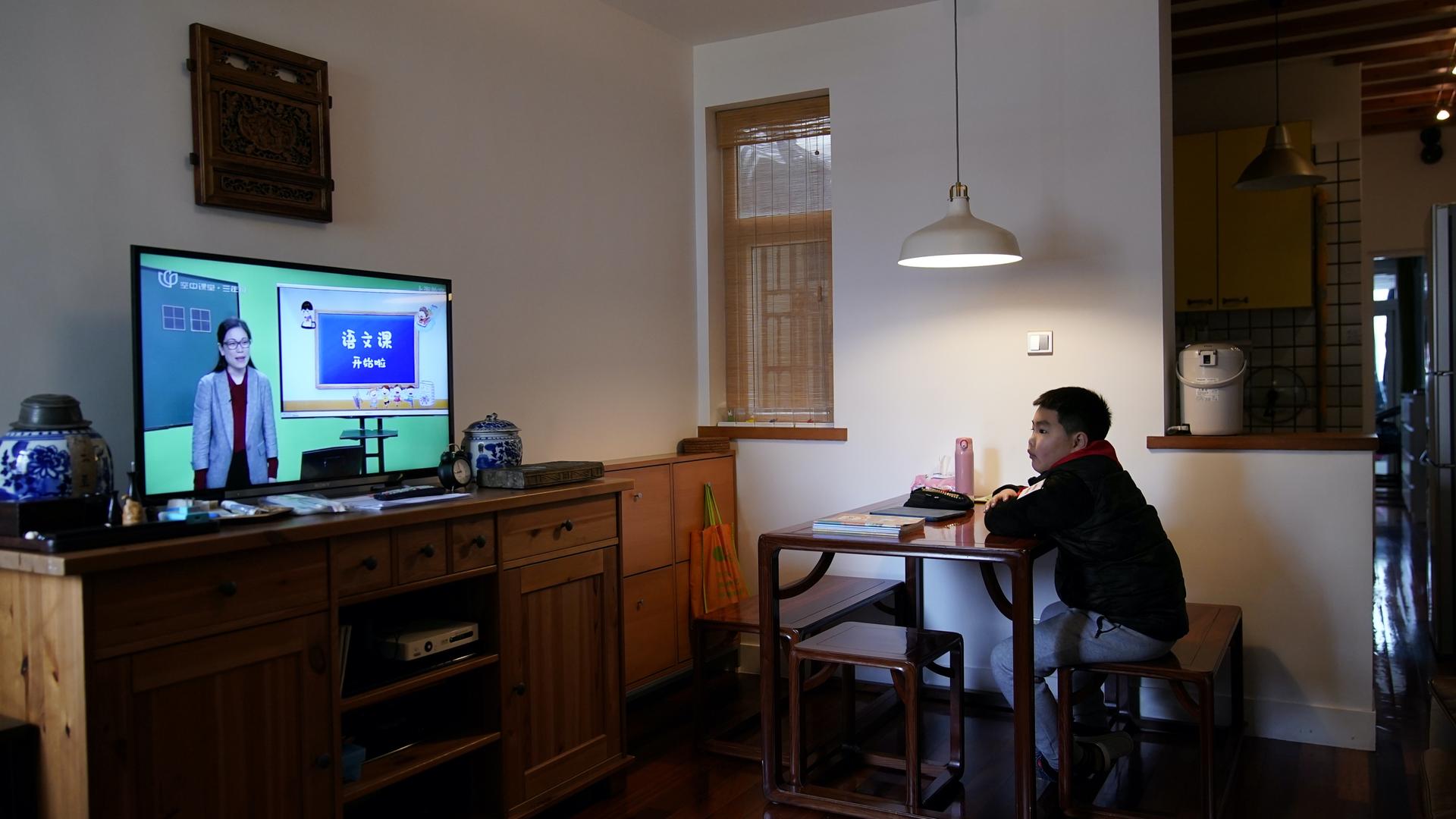 A boy sits in a living room at a desk and watches a TV with a lesson airing on it. 