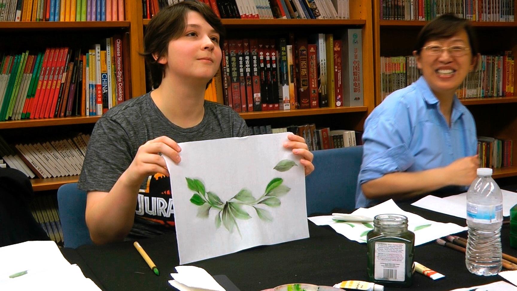 A white woman holds up a drawing of a plant next to a Chinese woman sitting at a table together 
