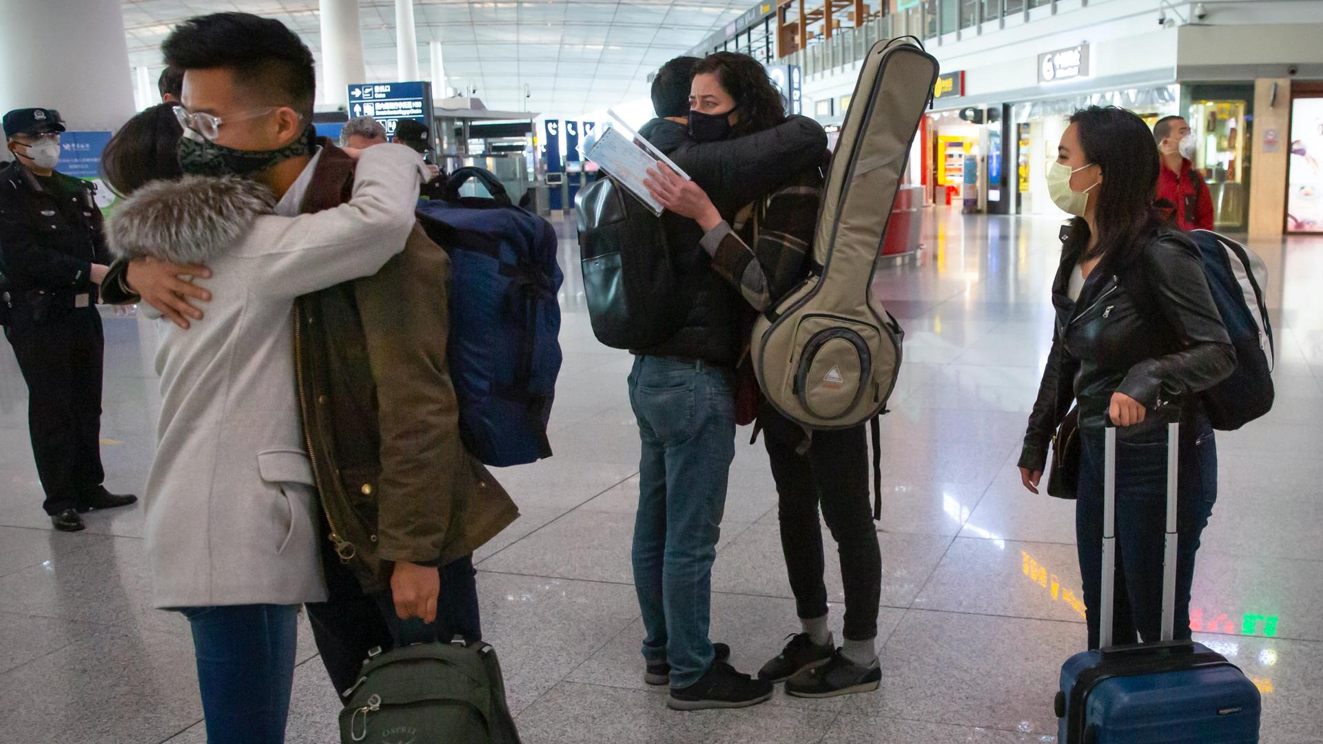 Pairs of individuals hug each other at an airport with their luggage on their shoulders and near their bodies 