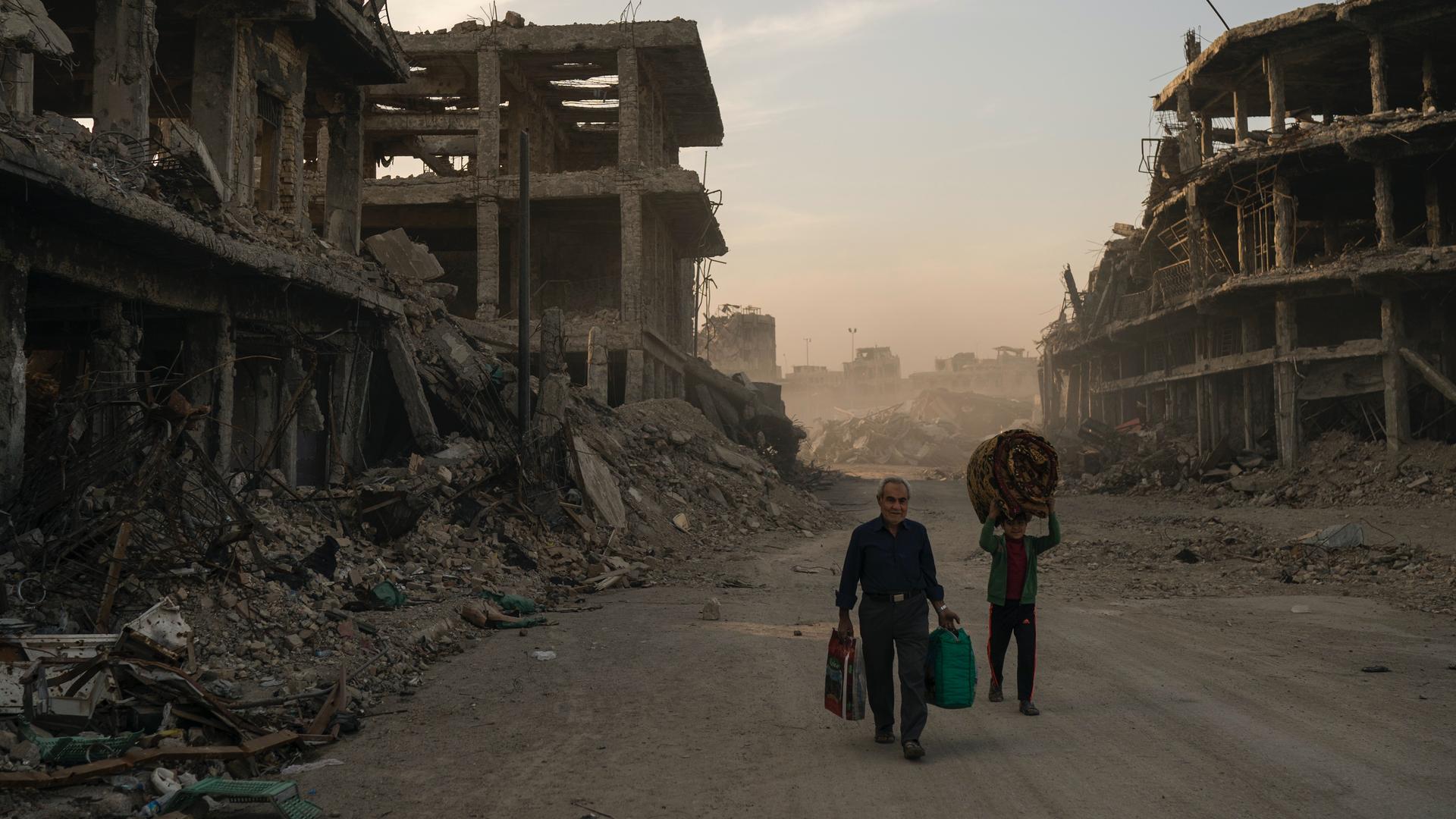 Two people carry luggage down a bombed out street in Mosul. 