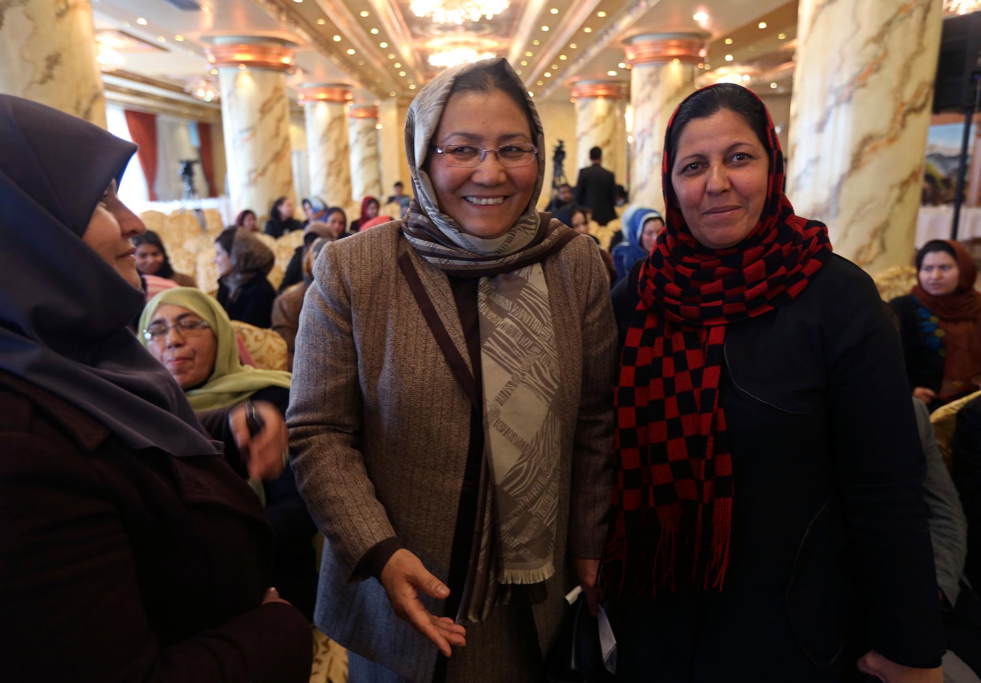 Three women wearing headscarves stand together and the one in the middle, wearing glasses, smiles.