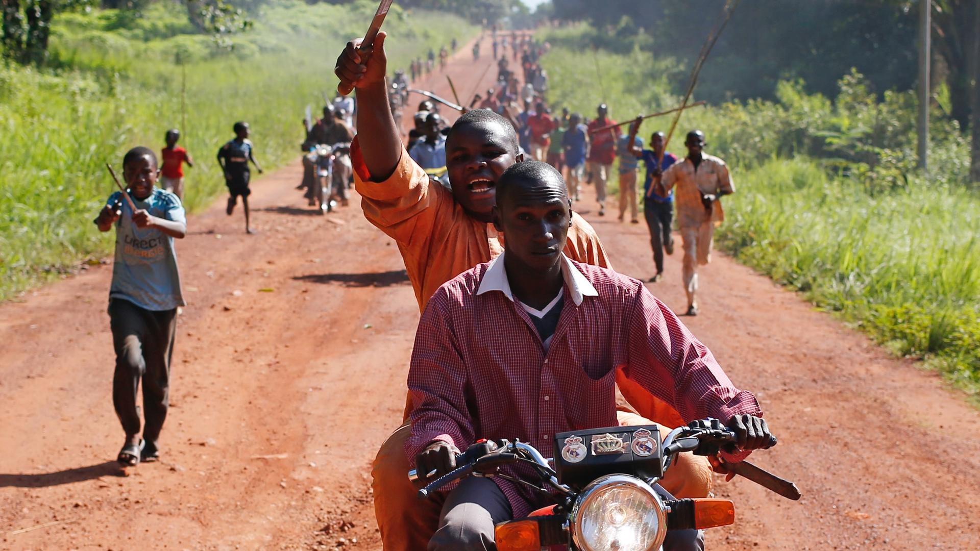 A man waves a machete in the air as he rides on the back of a motorcycle amid other protesters on foot on a long, dirt road. 
