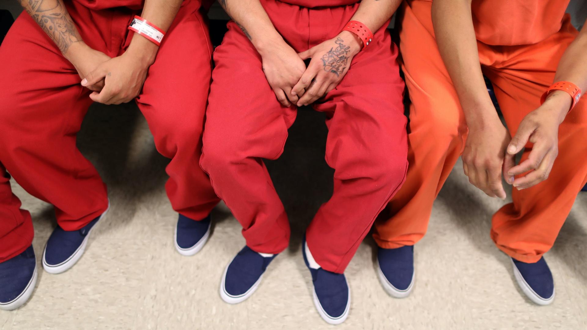 Three men wearing orange prison suits fold their hands in their laps and wear blue shoes. 