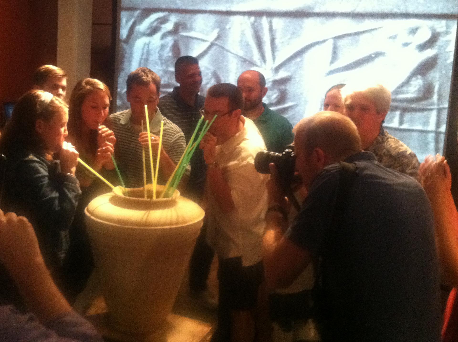 A group of people drink beer with straws out of a large vase.