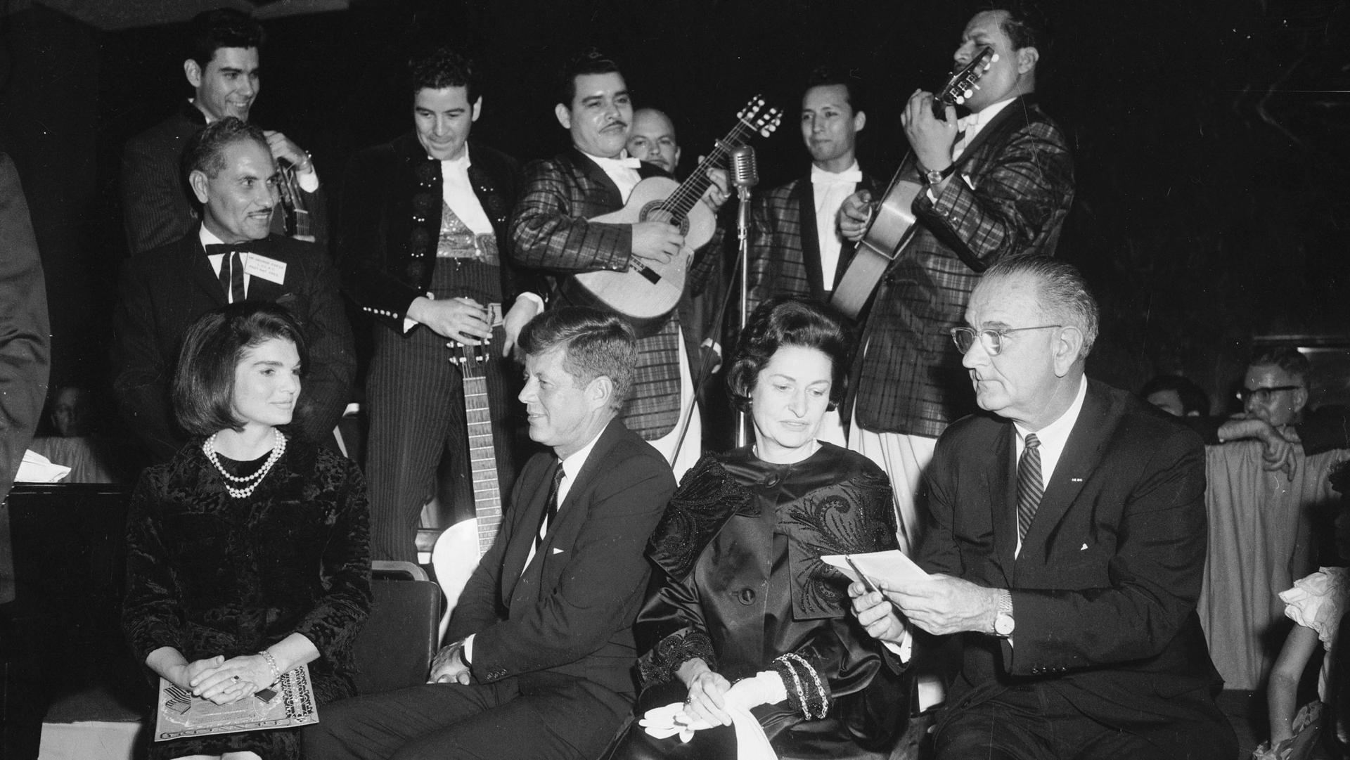 Foto en Blanco y negro del Presidente Kennedy y su esposa rodeados de musicos.