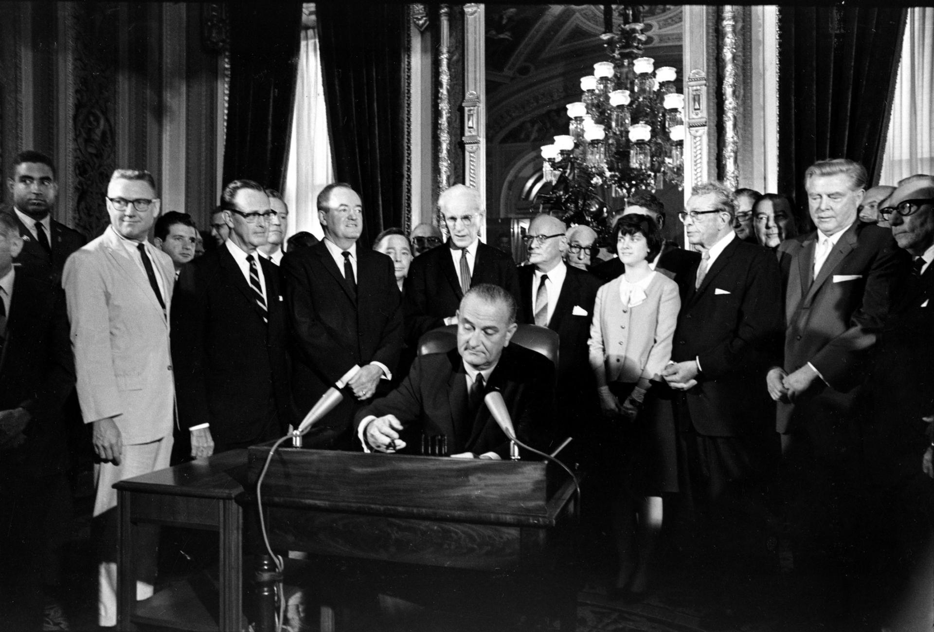 El Presidente Lyndon Johnson sentado en una mesa firmando un documento, aparece rodeado de personas.