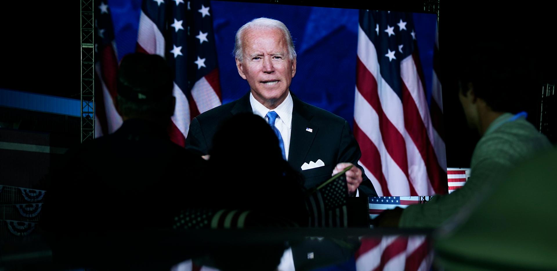 Joe Biden aparece en una pantalla mientras unas personas miran.