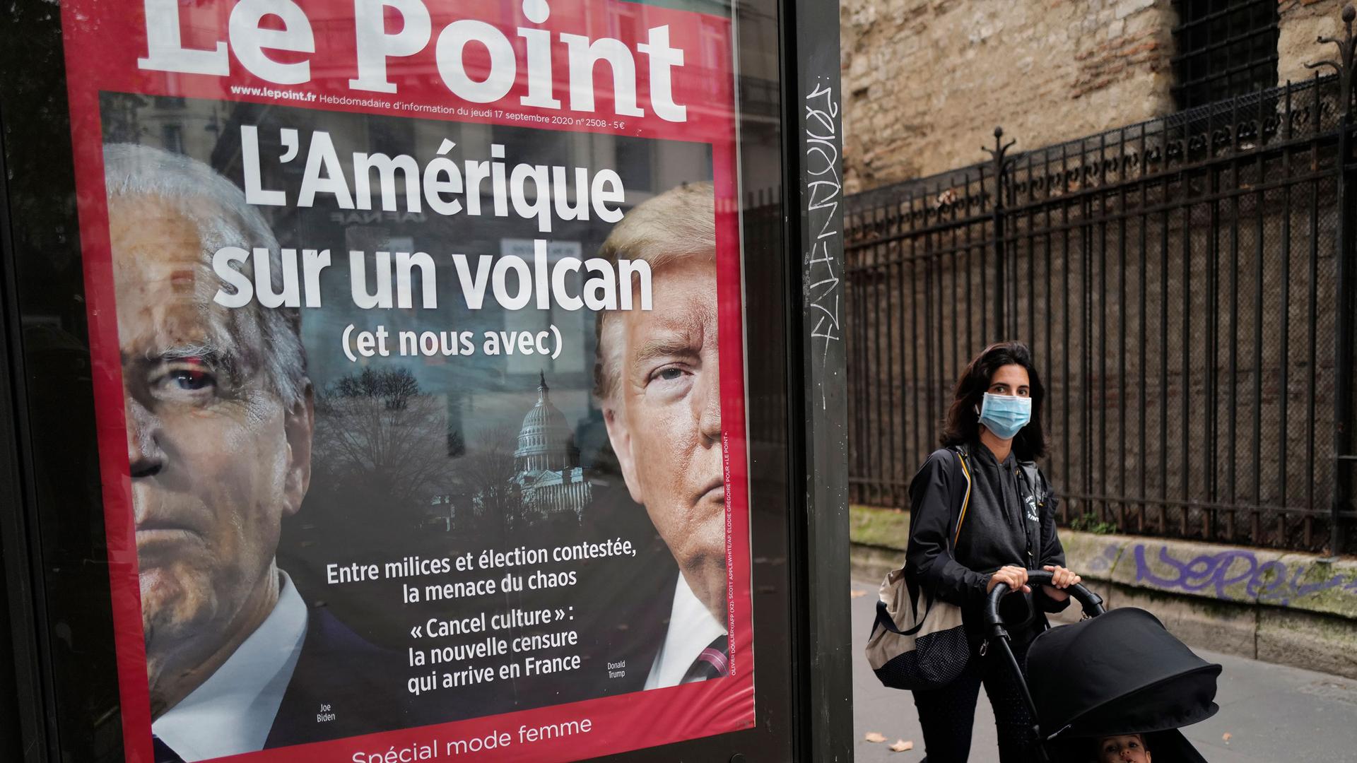 A small billboard for the French publication, Le Point, is show with the faces of Donald Trump and Joe Biden on it and a woman is shown walking past.