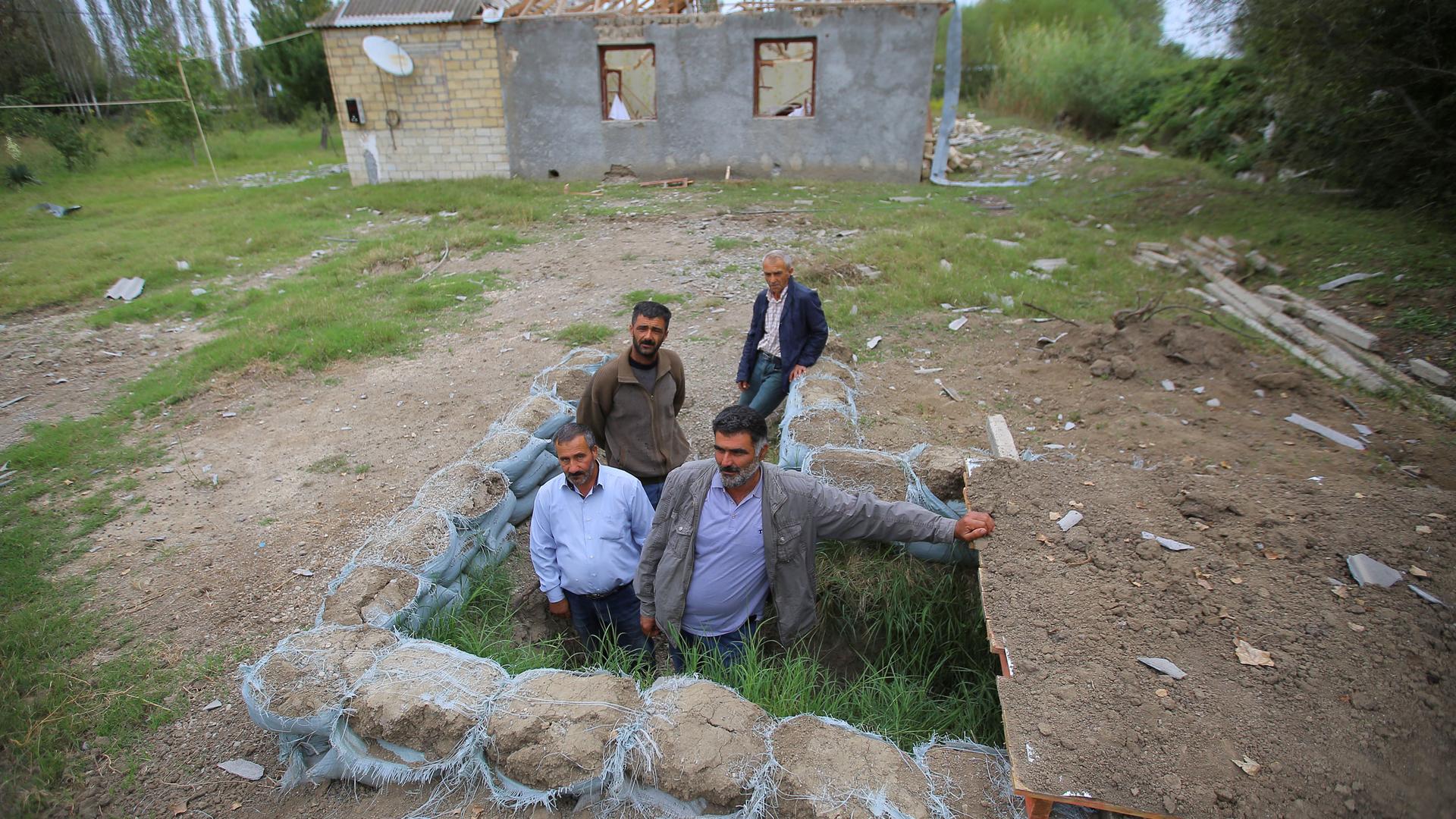Four men are shown standing in an dug out area lined with stone in an open area near a house.