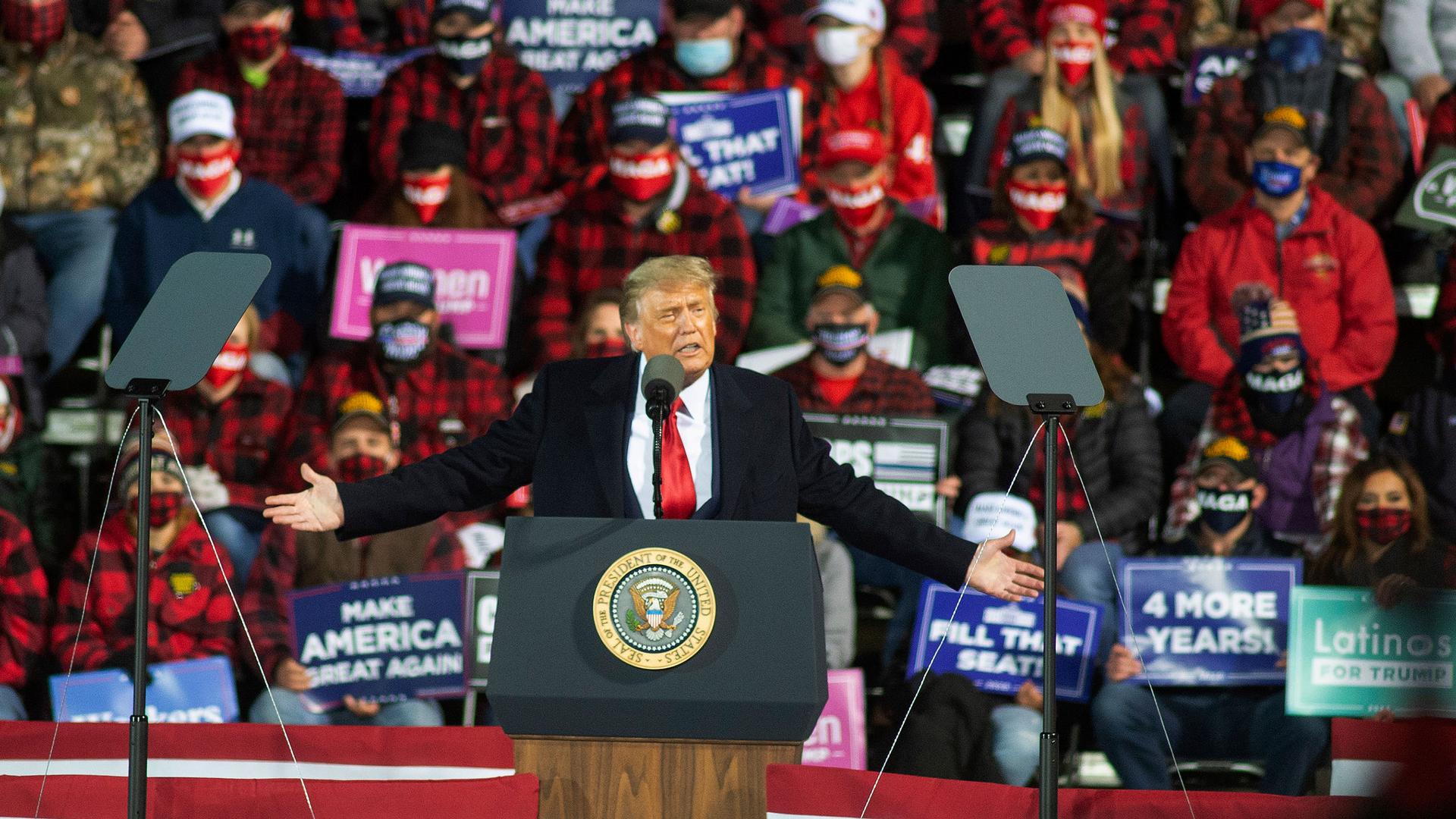 US President Donald Trump is shown with his arms outstretched, speaking at a podium with a crowd of people behind him.