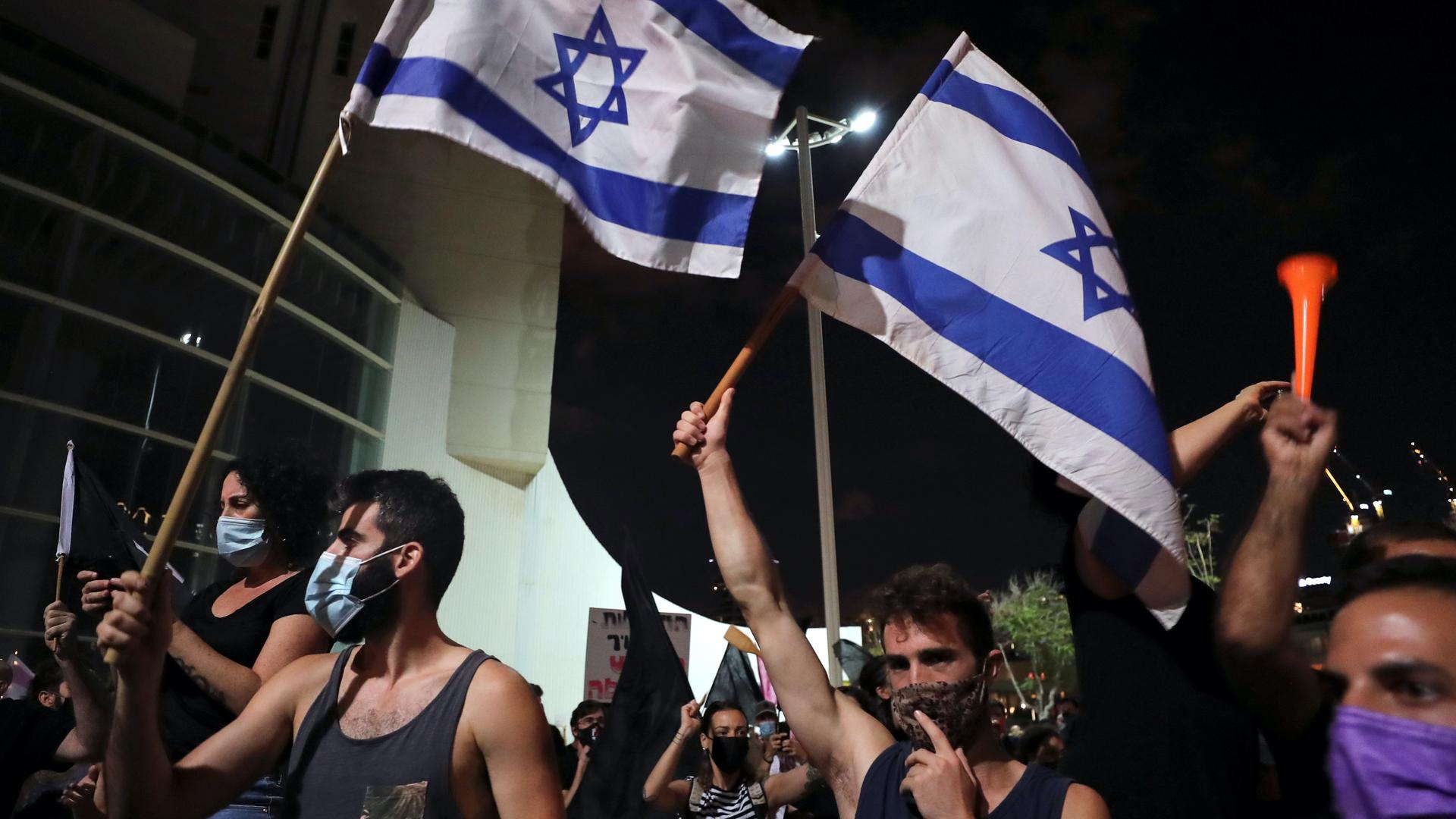 Two men wearing tank tops and face masks protest and wave blue and white Israeli flags in a big crowd