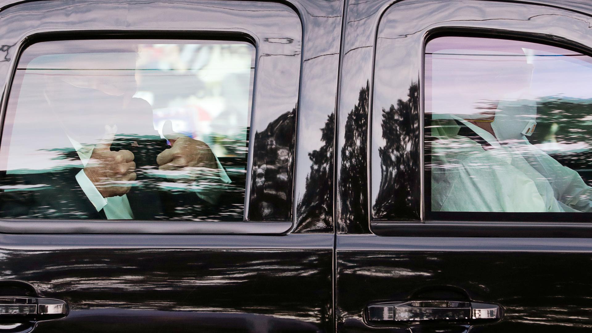 US President Donald Trump is shown through the glass of black SUV giving the thumbs up sign with both of his hands while wearing a face mask.