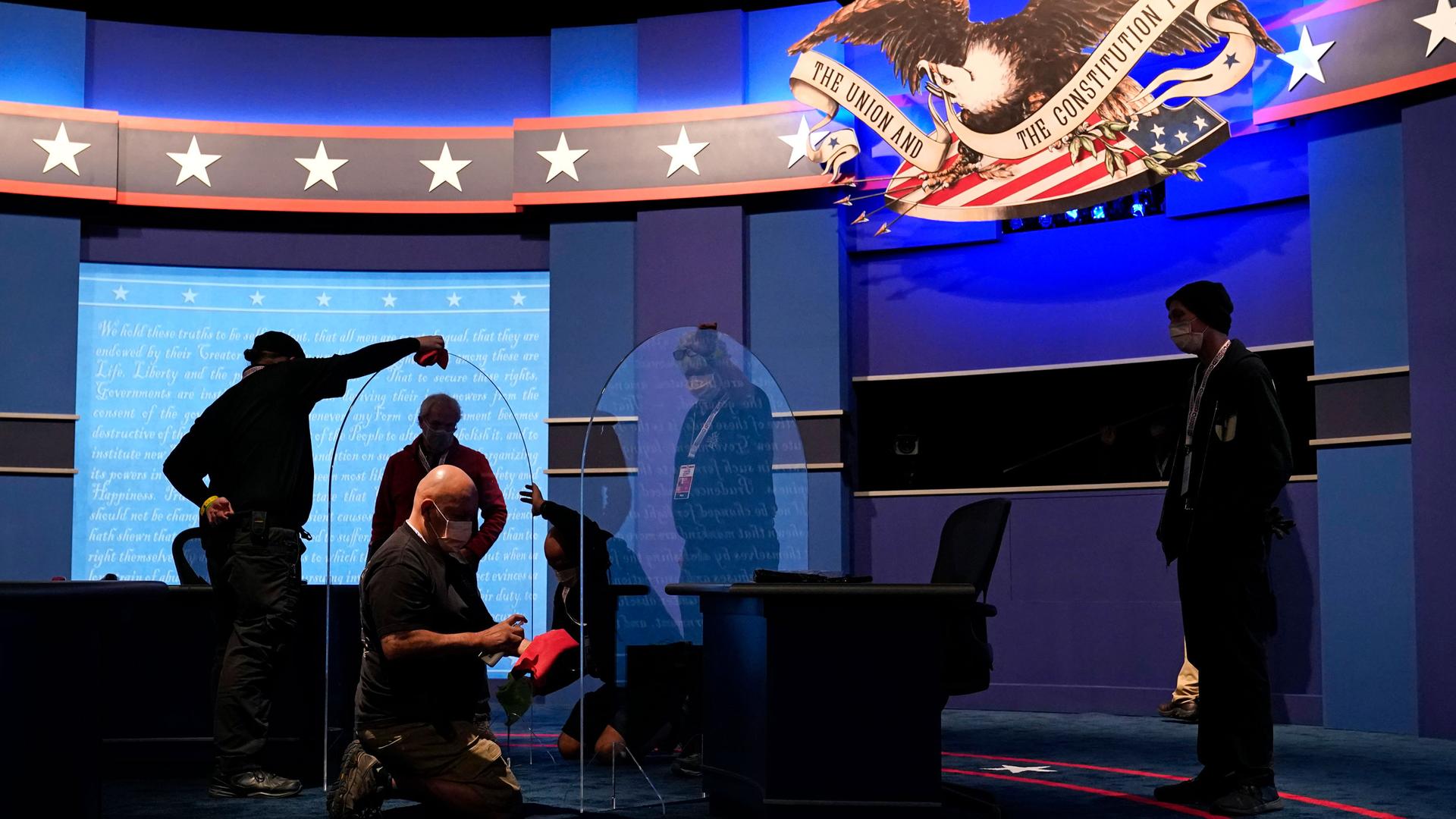 Workers clean protective plastic panels onstage between tables for Mike Pence and Kamala Harris with a large backdrop that includes stars across it.
