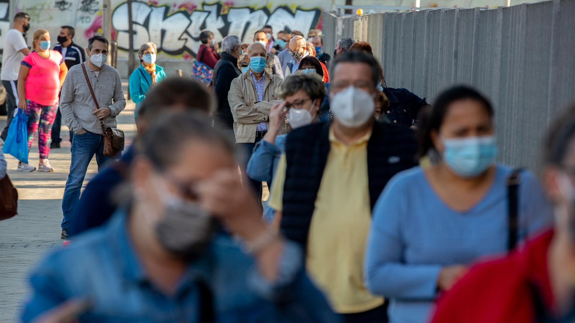 A long line of people are shown wearing face masks with wall painted with graffiti in the distance.