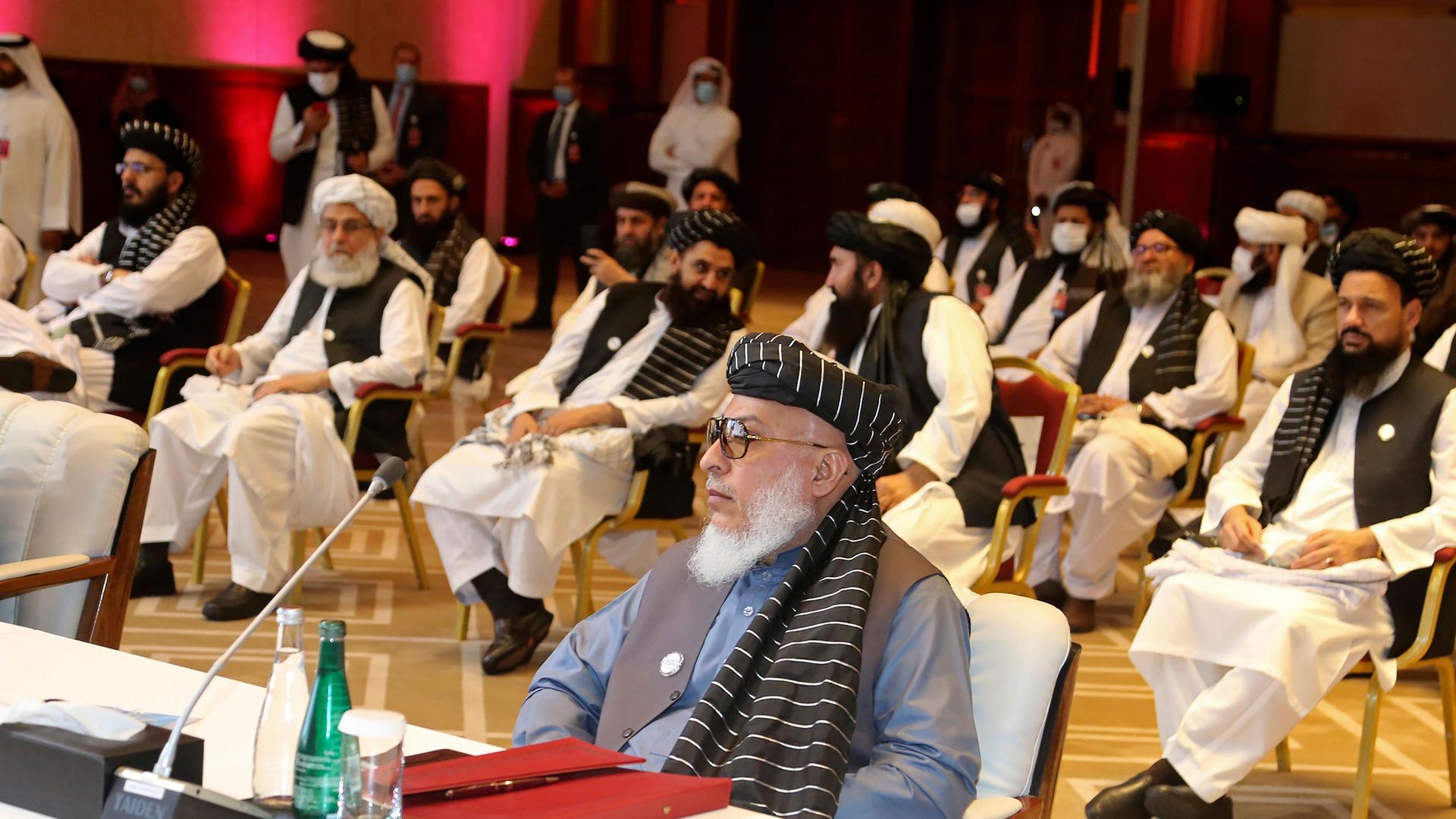 Several members of the Taliban negotiating delegation are shown seated in chairs spaced several feet from each other in a large room.
