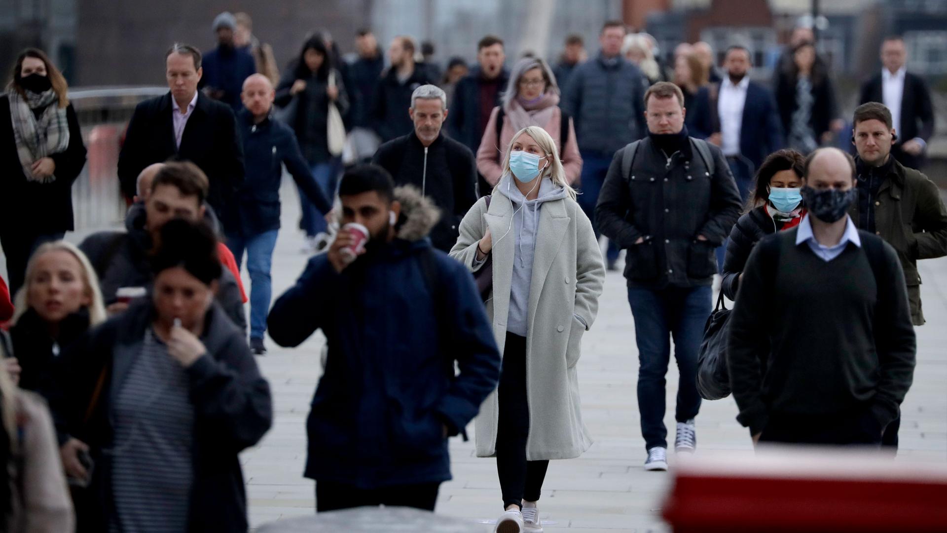 A crowd of people are shown walking across the London Bridge, many wearing face masks.