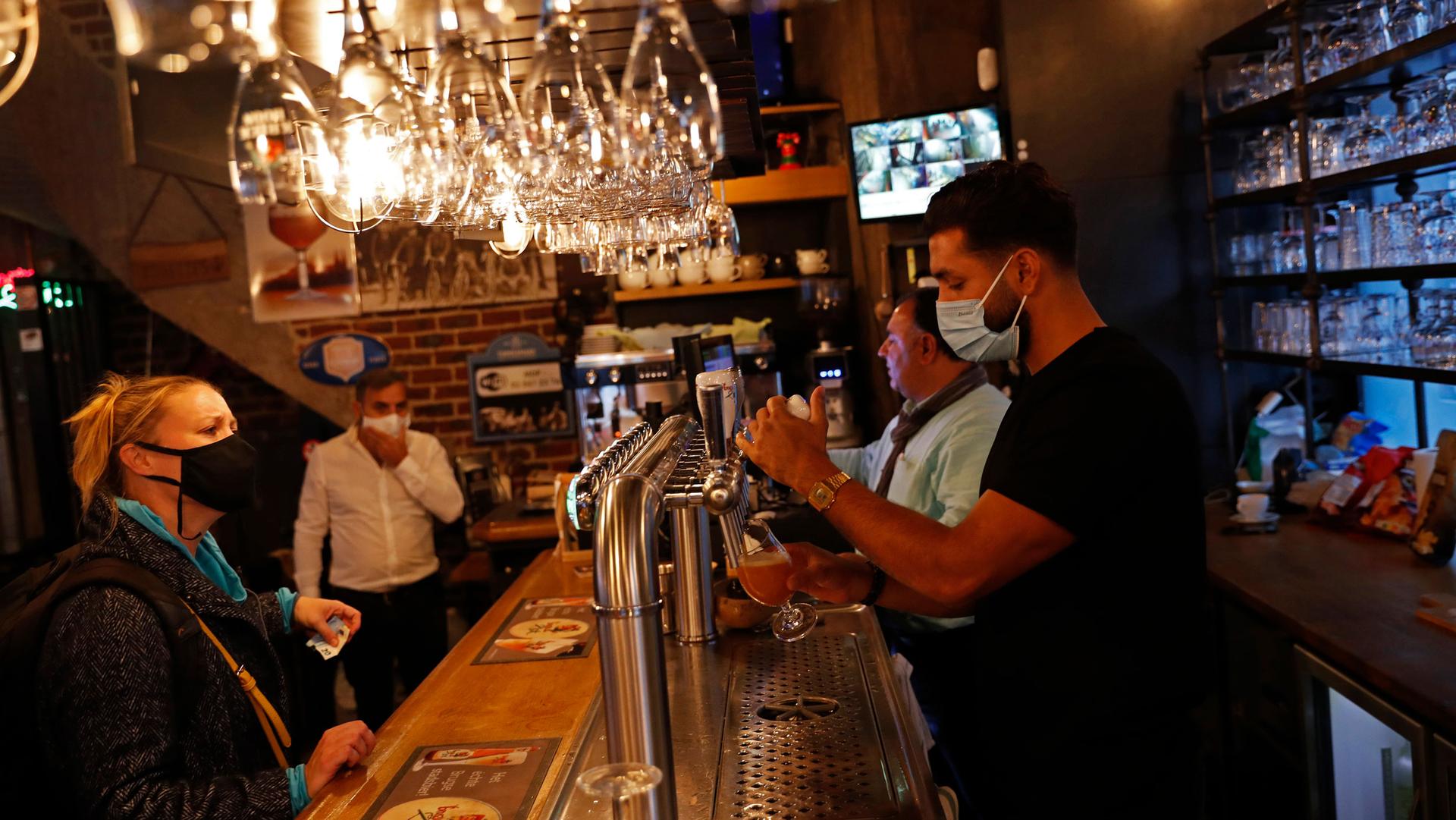 A bartender is shown pulling a draft beer while wearing a face maks with glasses hanging above the bar.