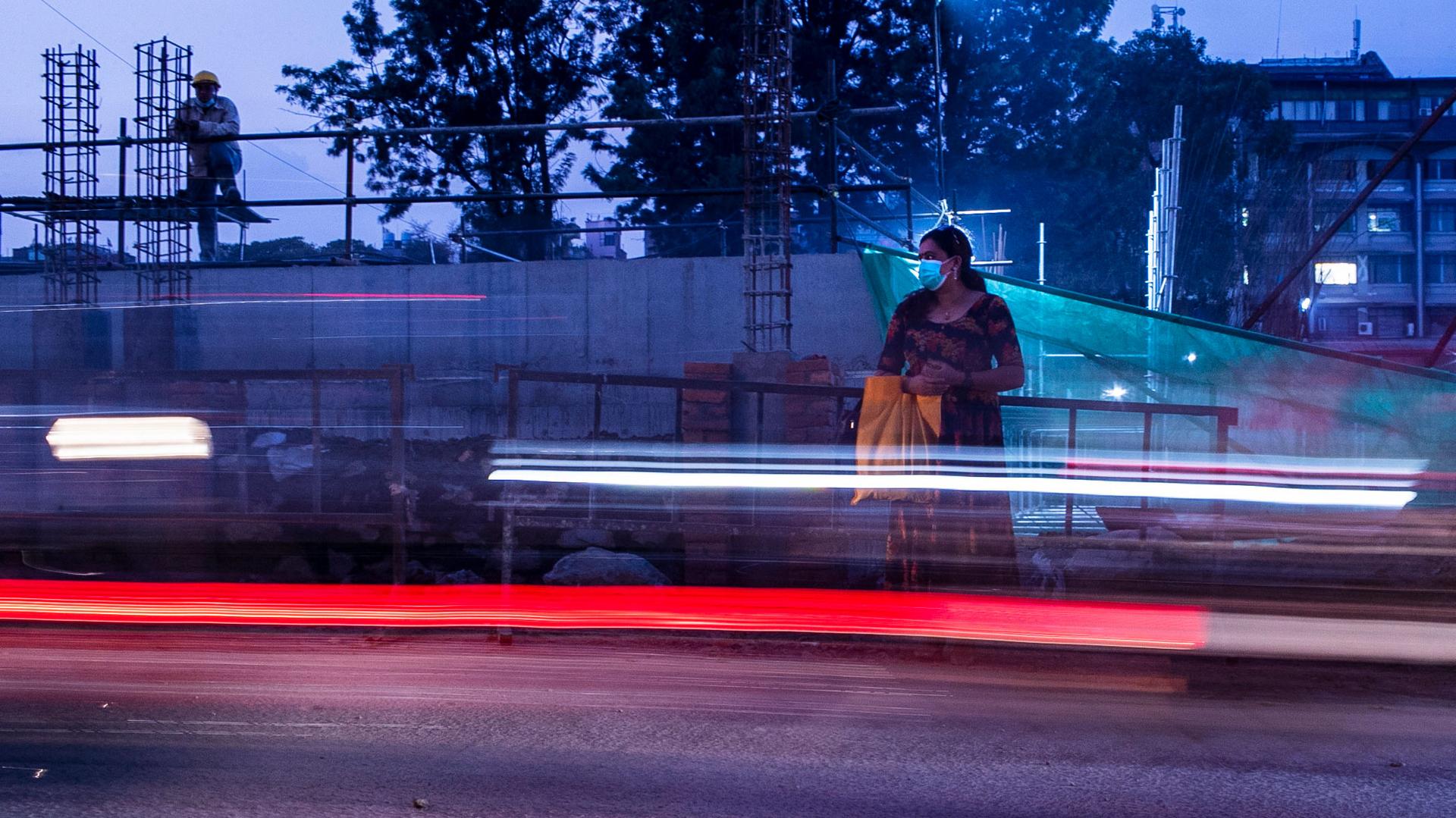 A womanis shown holding a bag and wearing a face mask with a streak of the bright colors of a car blurred as it passes by.