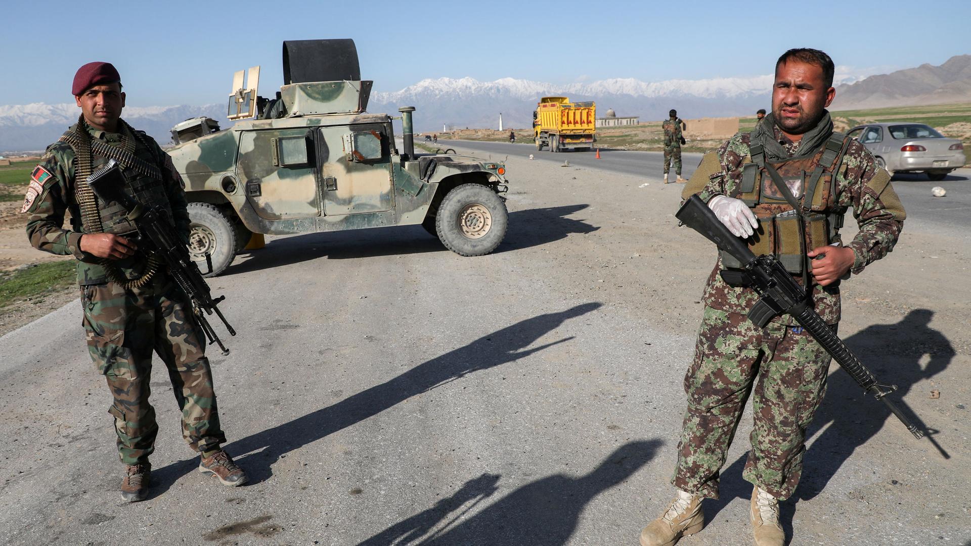 Two soldiers are shown standing near a road and holding weapons with an armored vehicle parked behind them.