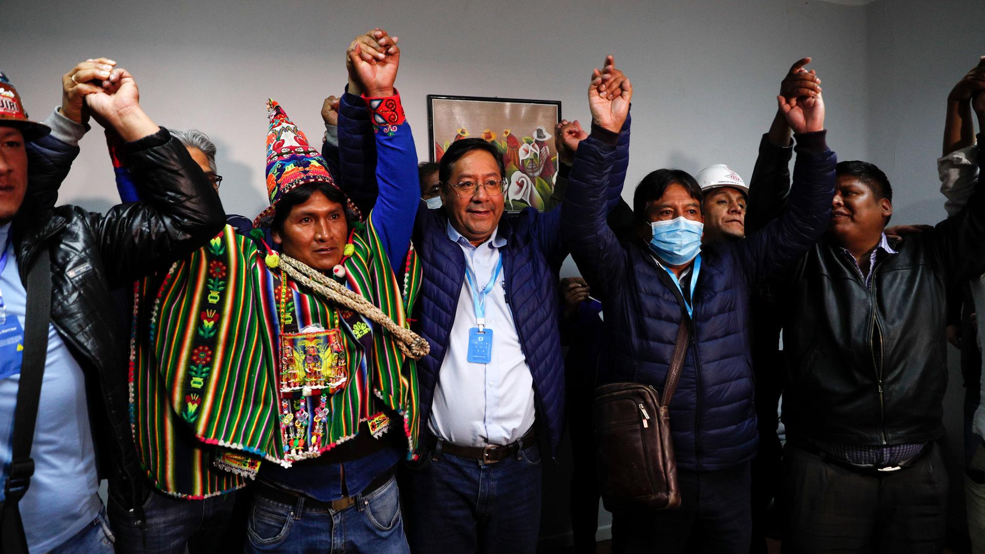 Several Bolivian politicians are shown in a room holding their hands in the air in celebration.