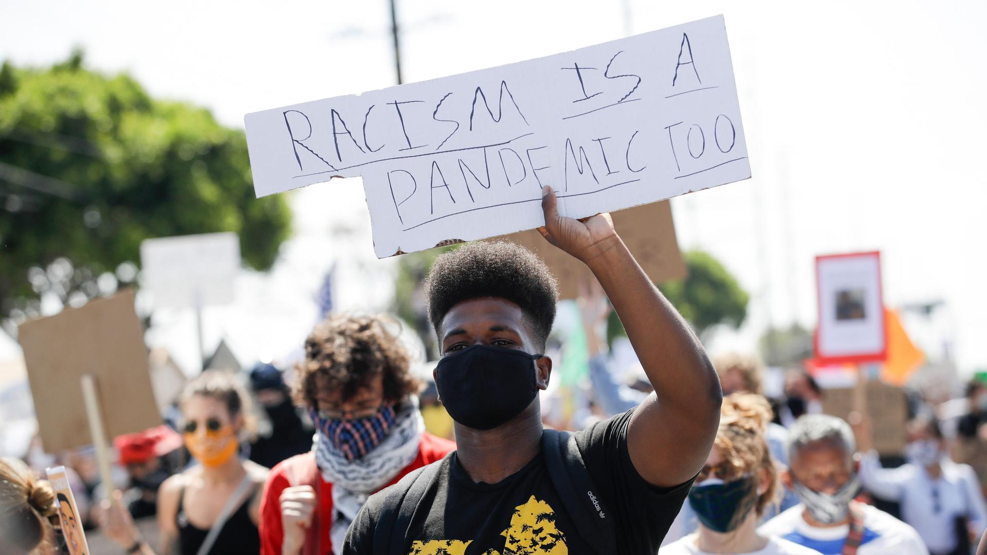 A crowd of protesters are shown with a man in the center holding a sign that reads, "Racism is a pandemic too."