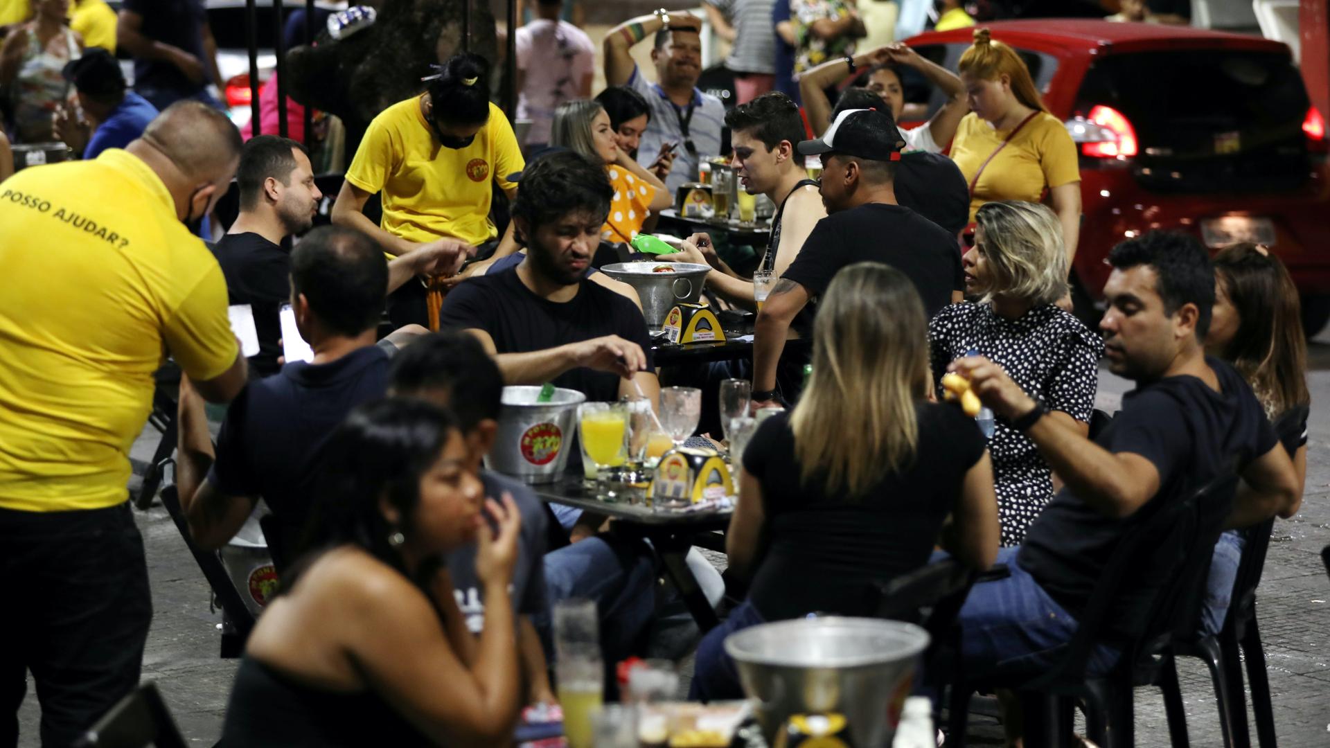 People enjoy beers and food at an outdoor restaurant where waitstaff wear yellow shirts. 