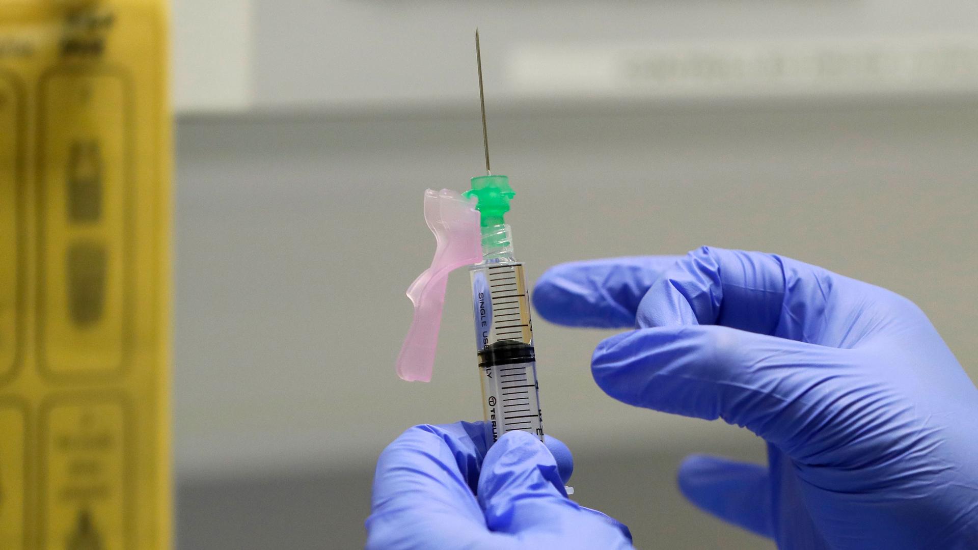 A close-up photo of a researcher's gloved hands and a syringe filled with a vaccine trial.