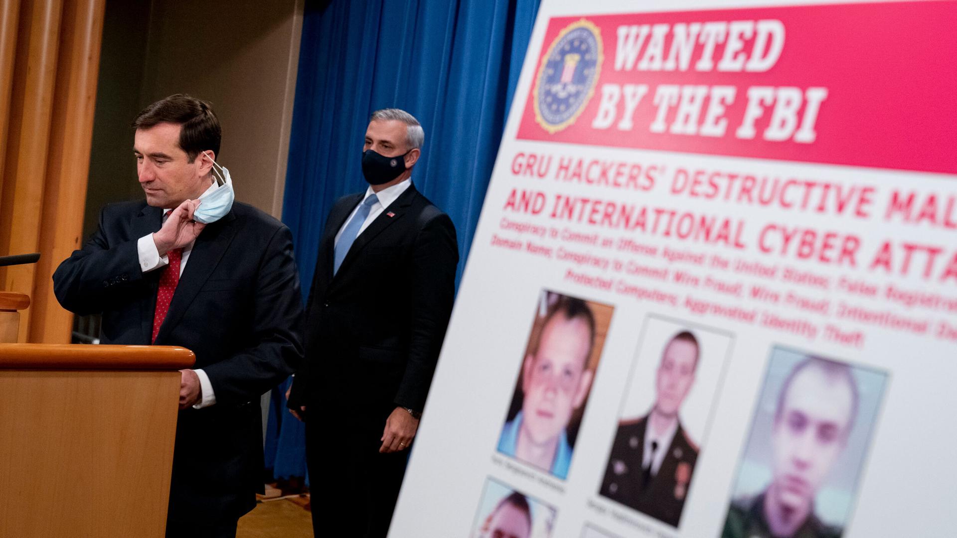 A poster showing six wanted Russian military intelligence officers is displayed as Assistant Attorney General for the National Security Division John Demers, left, takes the podium to speak at a news conference at the Department of Justice, Oct. 19, 2020.