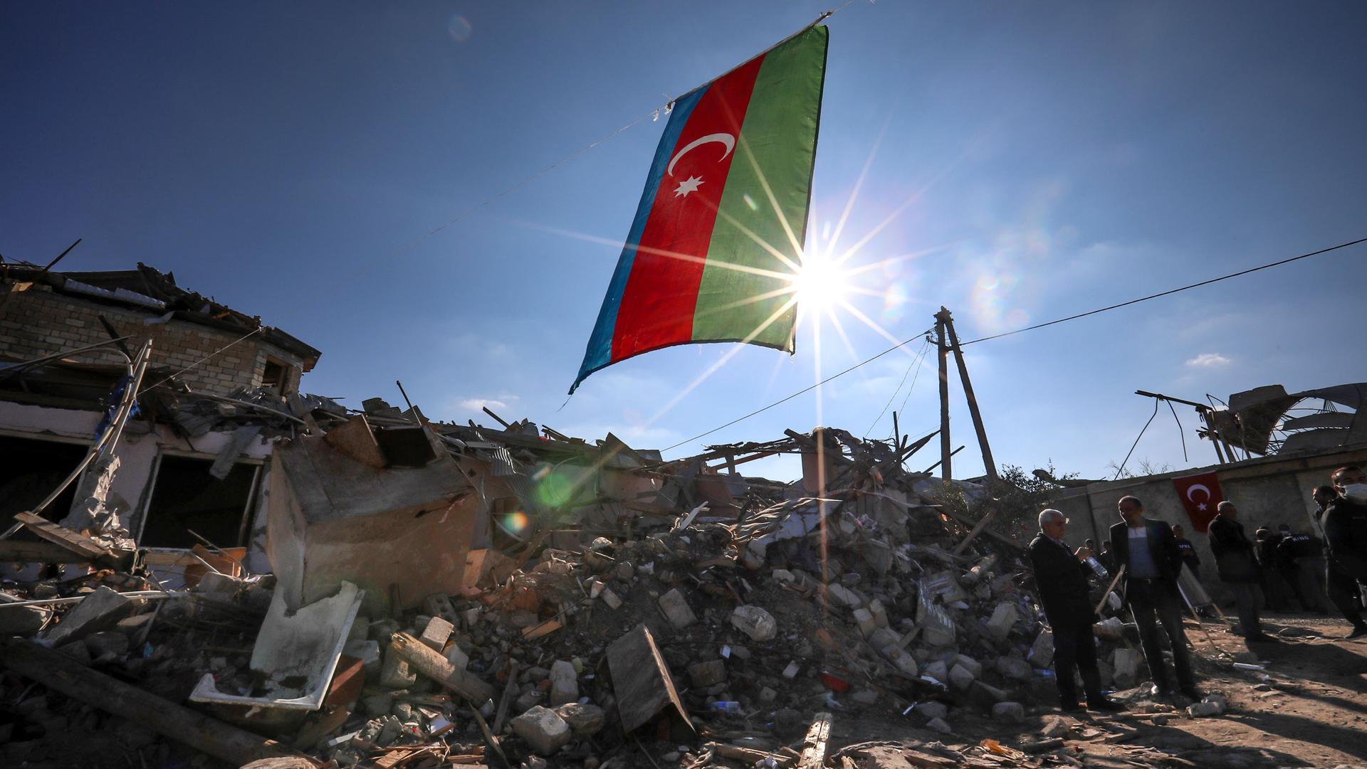 Azerbaijan's national flag is shown flying over destroyed houses with bright rays of sunlight backlighting it.