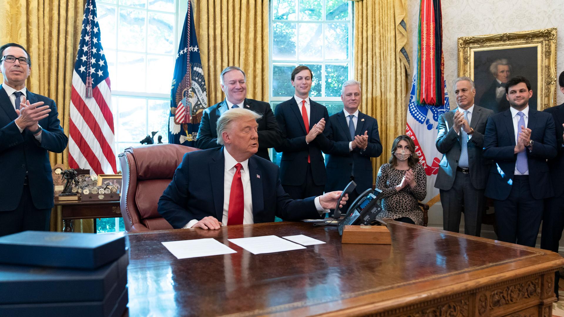 Donald Trump is shown sitting at a desk and wearing a dark suit with a red tie and hanging up a phone.