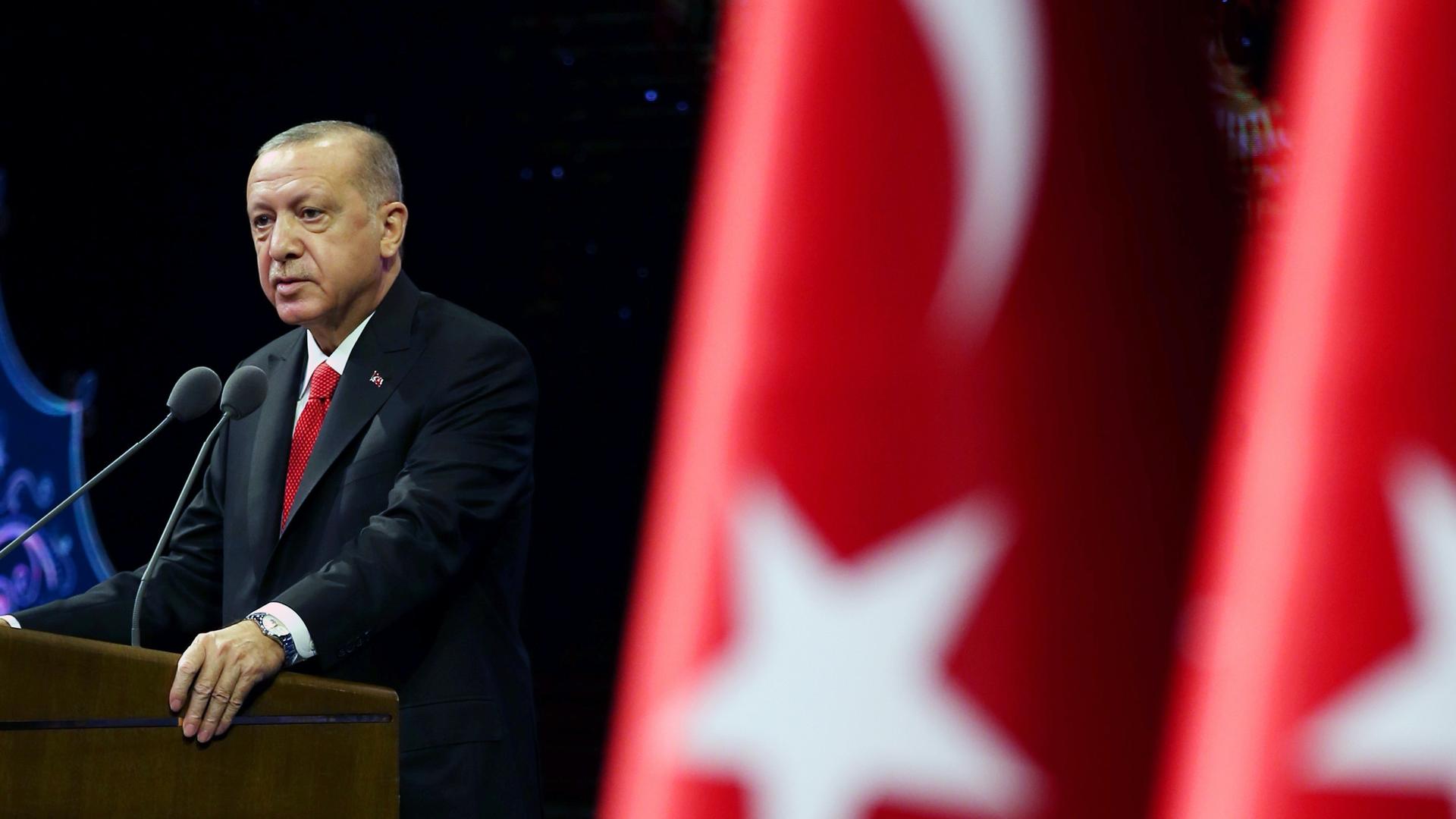 A man in a suit stands at a podium near red and white Turkish flag. 