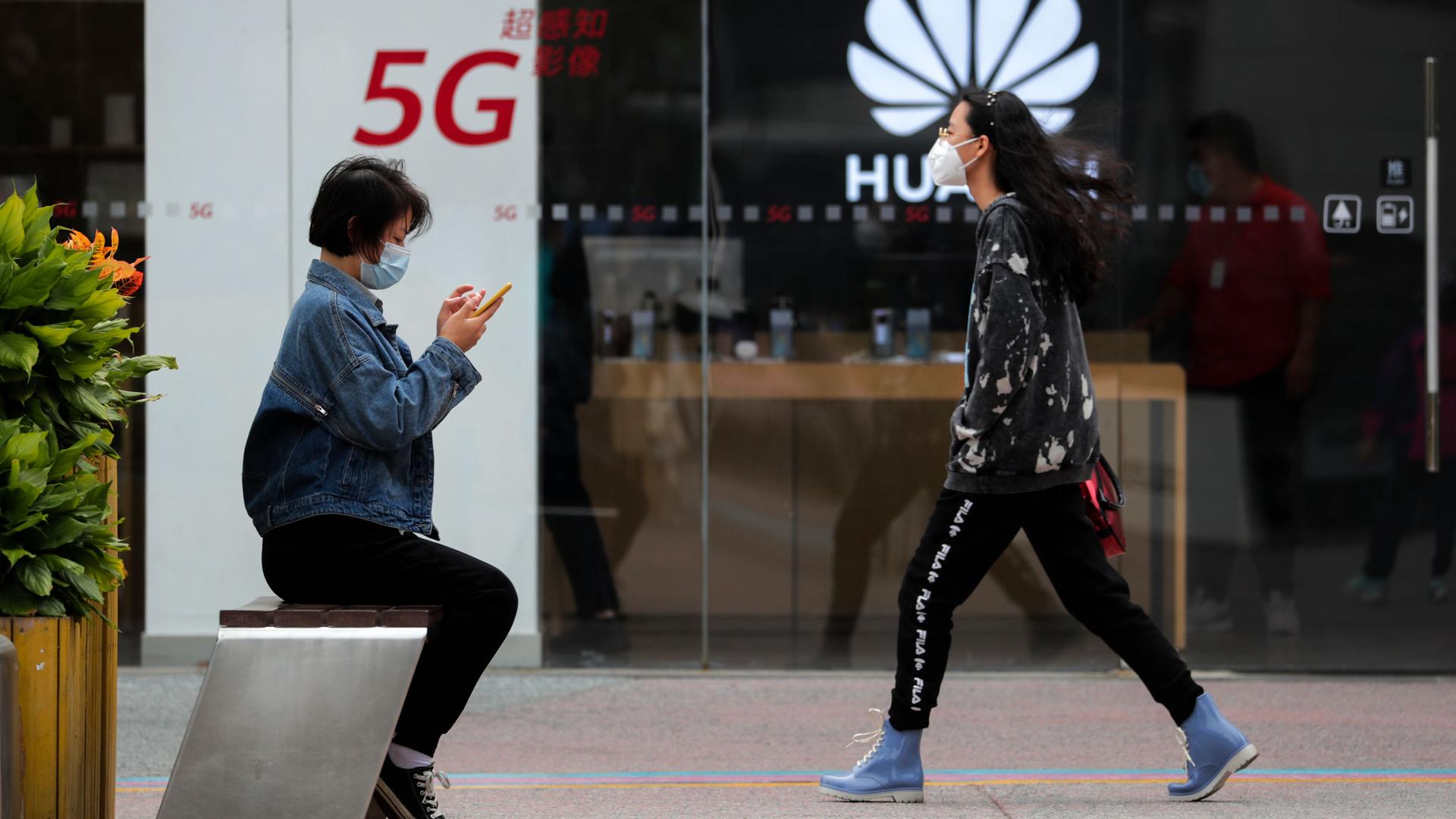A woman wearing a jean jacket is shown sitting on a bench as anothe woman walks toward her with the clear glass of a Huawei store in the distance.