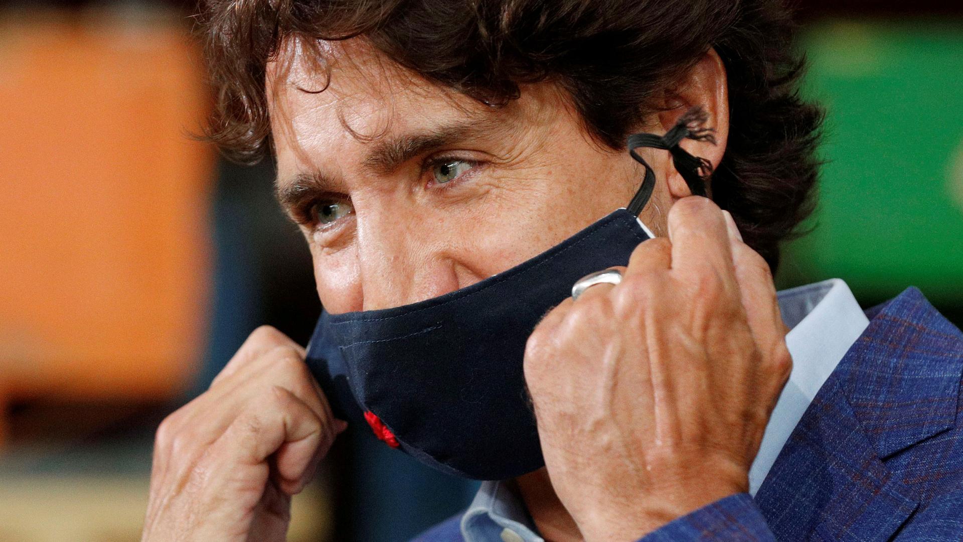 Canada's Prime Minister Justin Trudeau is shown in a close-up photo removing a dark face mask and wearing plaid suit coat.