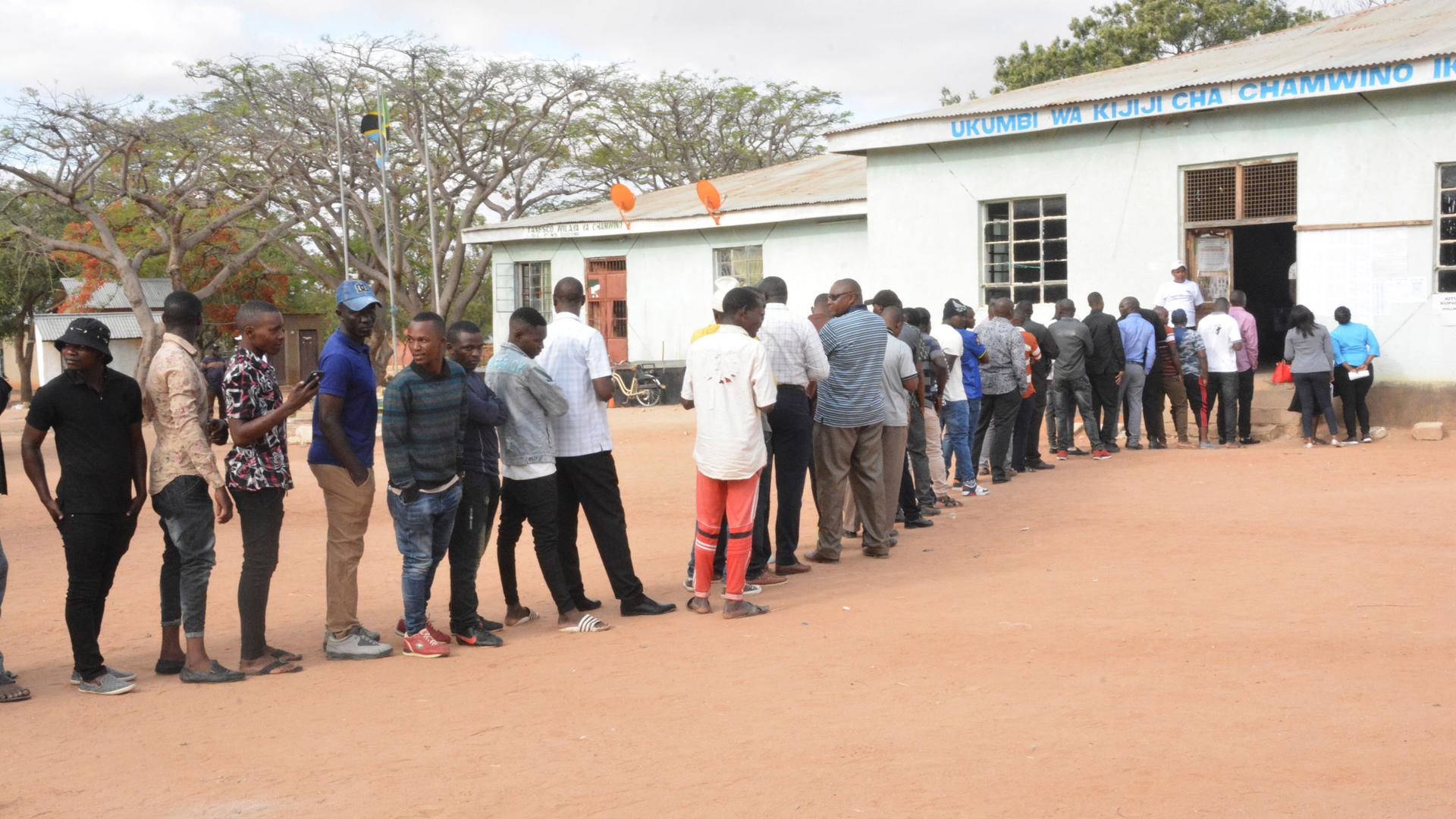 A long line of people are shown standing outside of a polling location in Tanzania.