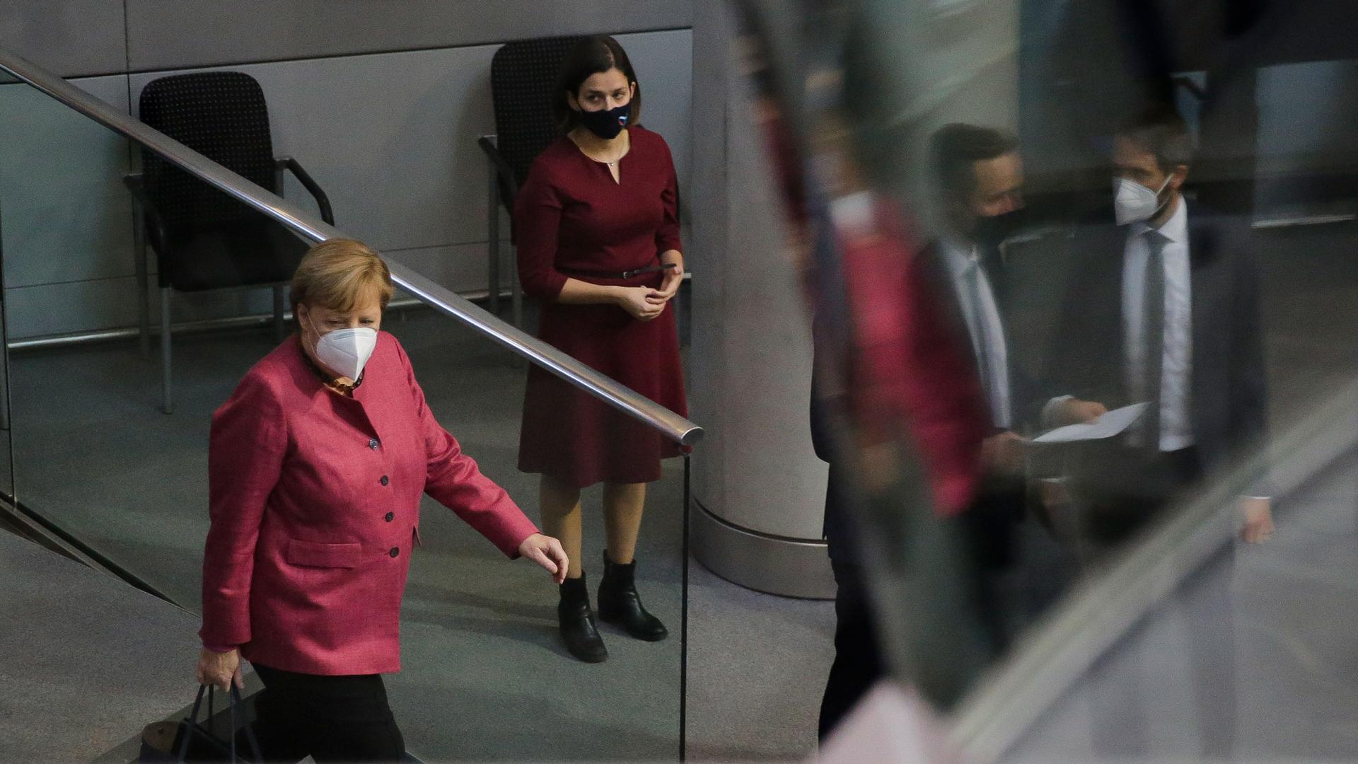 German Chancellor Angela Merkel is shown walking with a briefcase and wearing a red jacket with a woman behind her wearing a dark red dress.