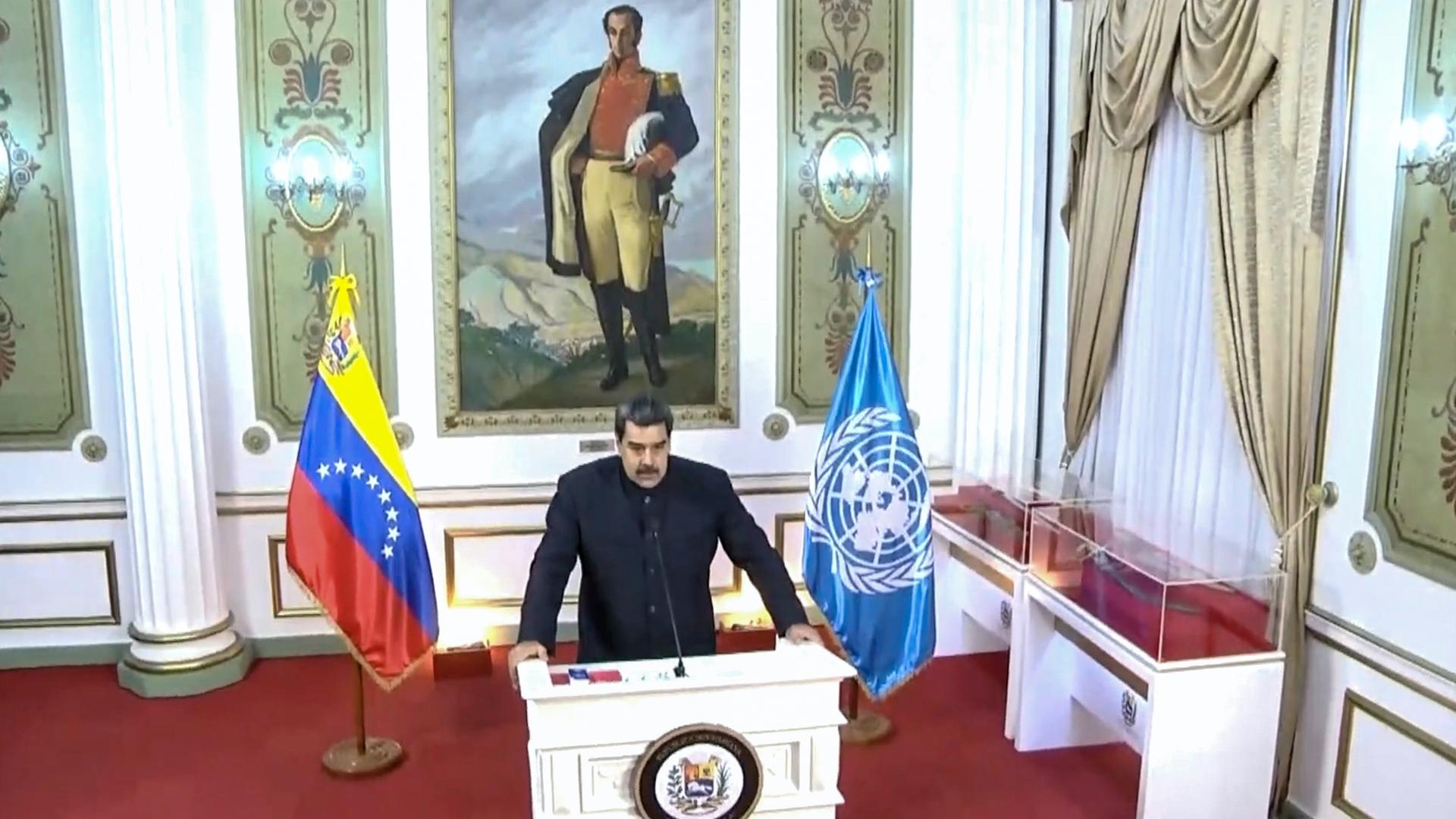 Maduro speaks at a white podium flanked by two flags in a decorative room with a painting of a uniformed man behind him. 