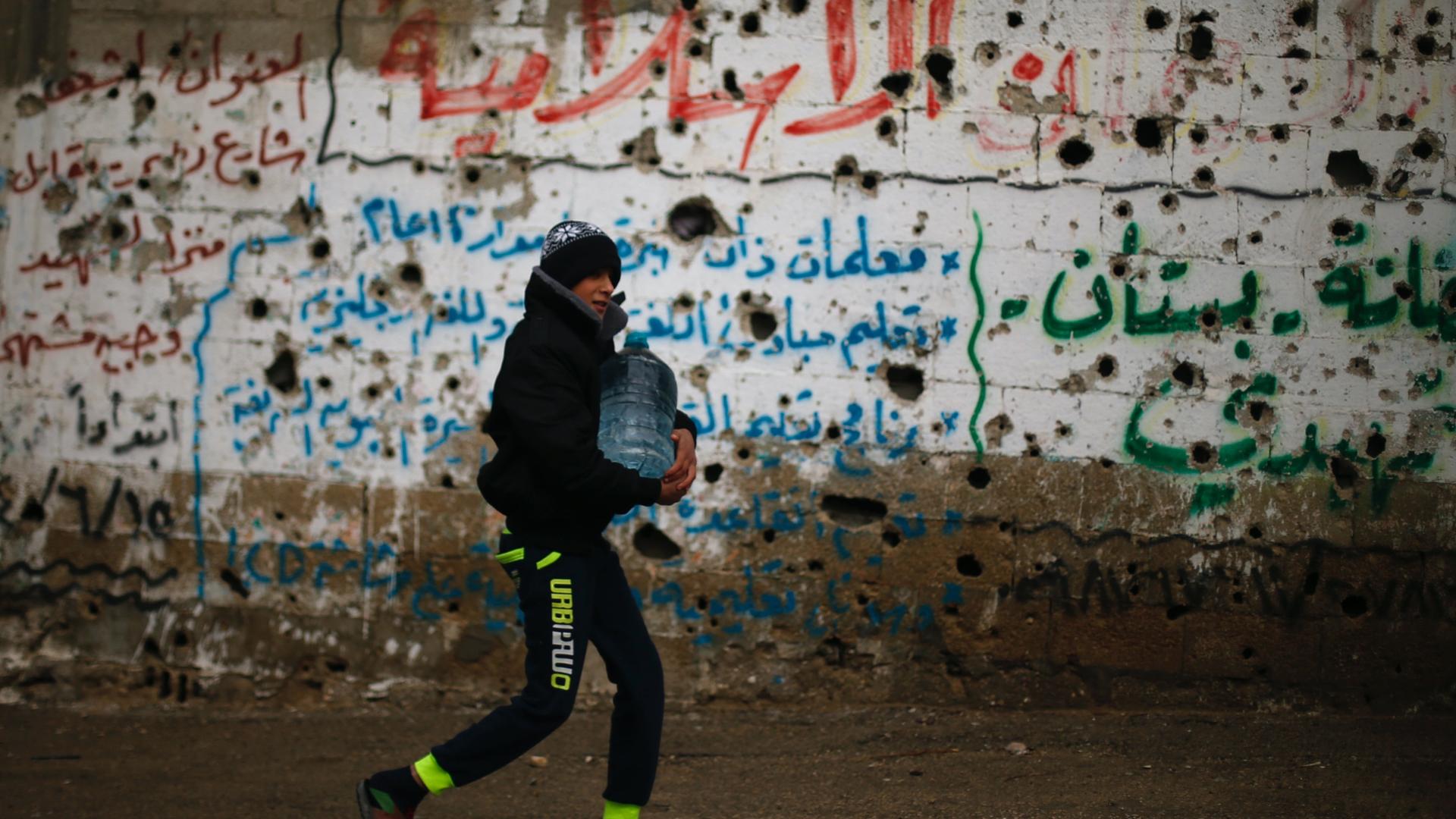 A boy carries a tub of water down a street past a wall full of grafitti in Arabic letters. 