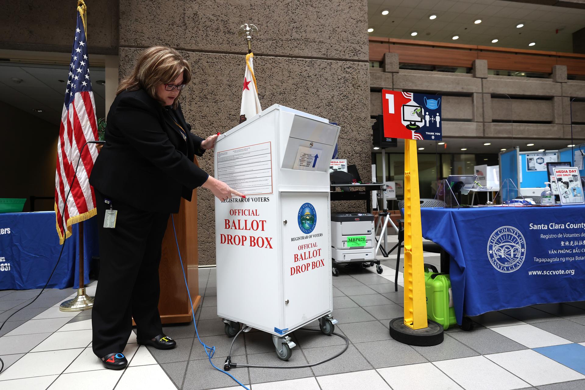 A woman stands next to a ballot box, pointing at specific details.