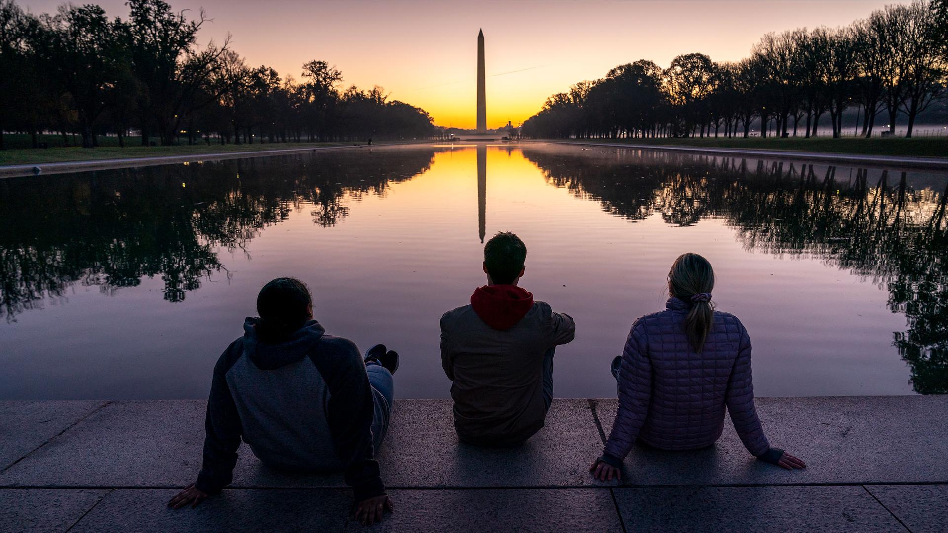 Three people are shown sitting on the ground in front of a long reflecting pool with the Washington Monument in the distance as the sun comes up.