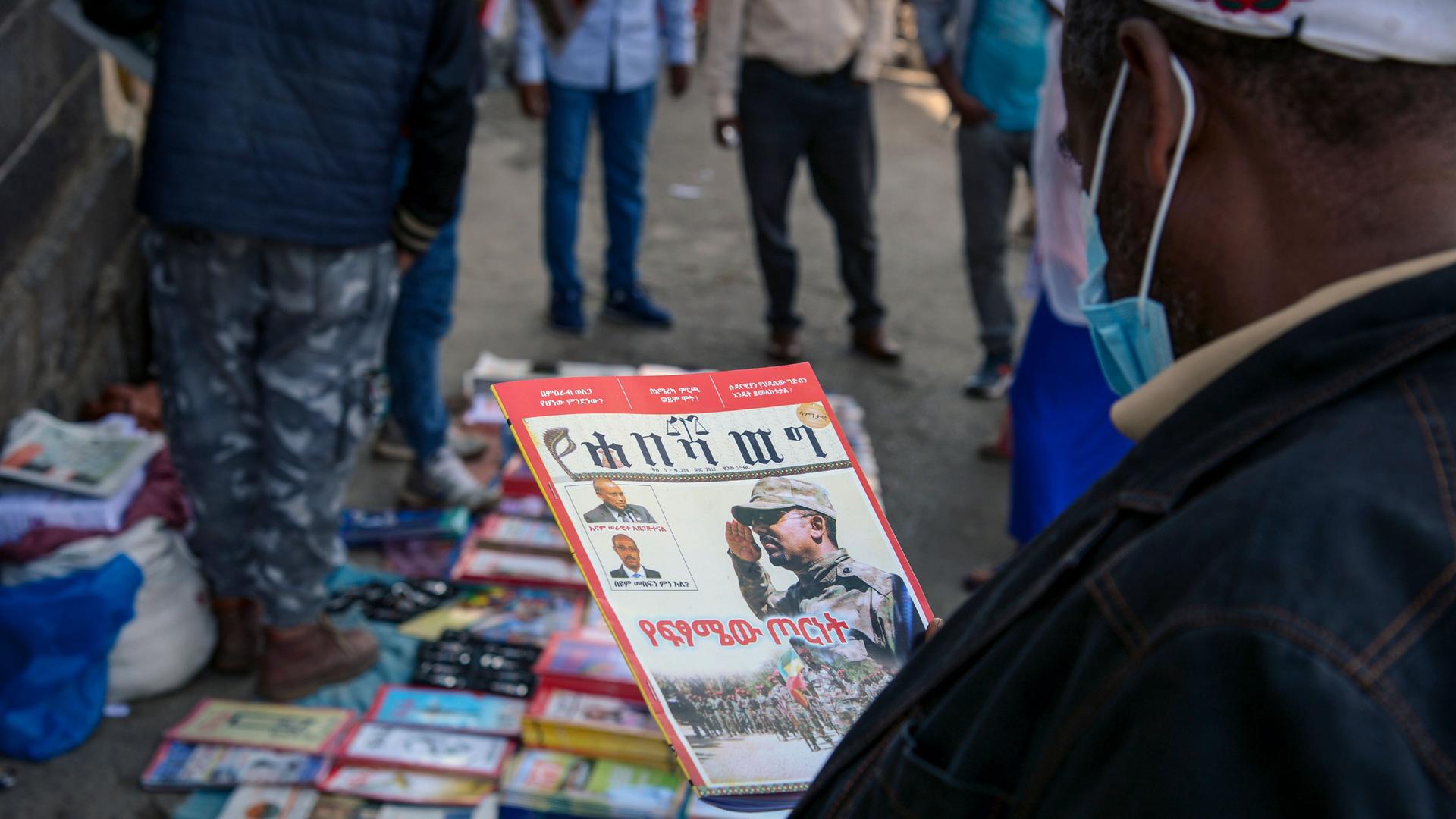A man is shown wearing a face mask and holding a magazine with dozens of other magazines for sale and set on the ground.