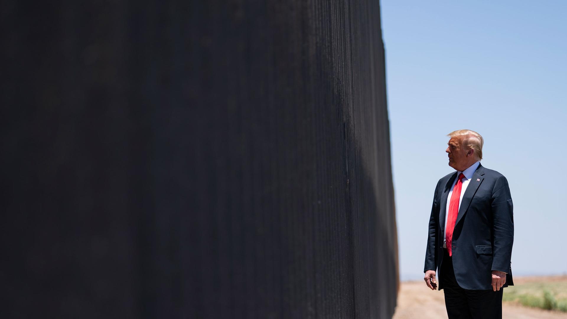 President Donald Trump is shown wearing a blue suit and red tie while looking at the dark facade of the US-Mexico border wall.
