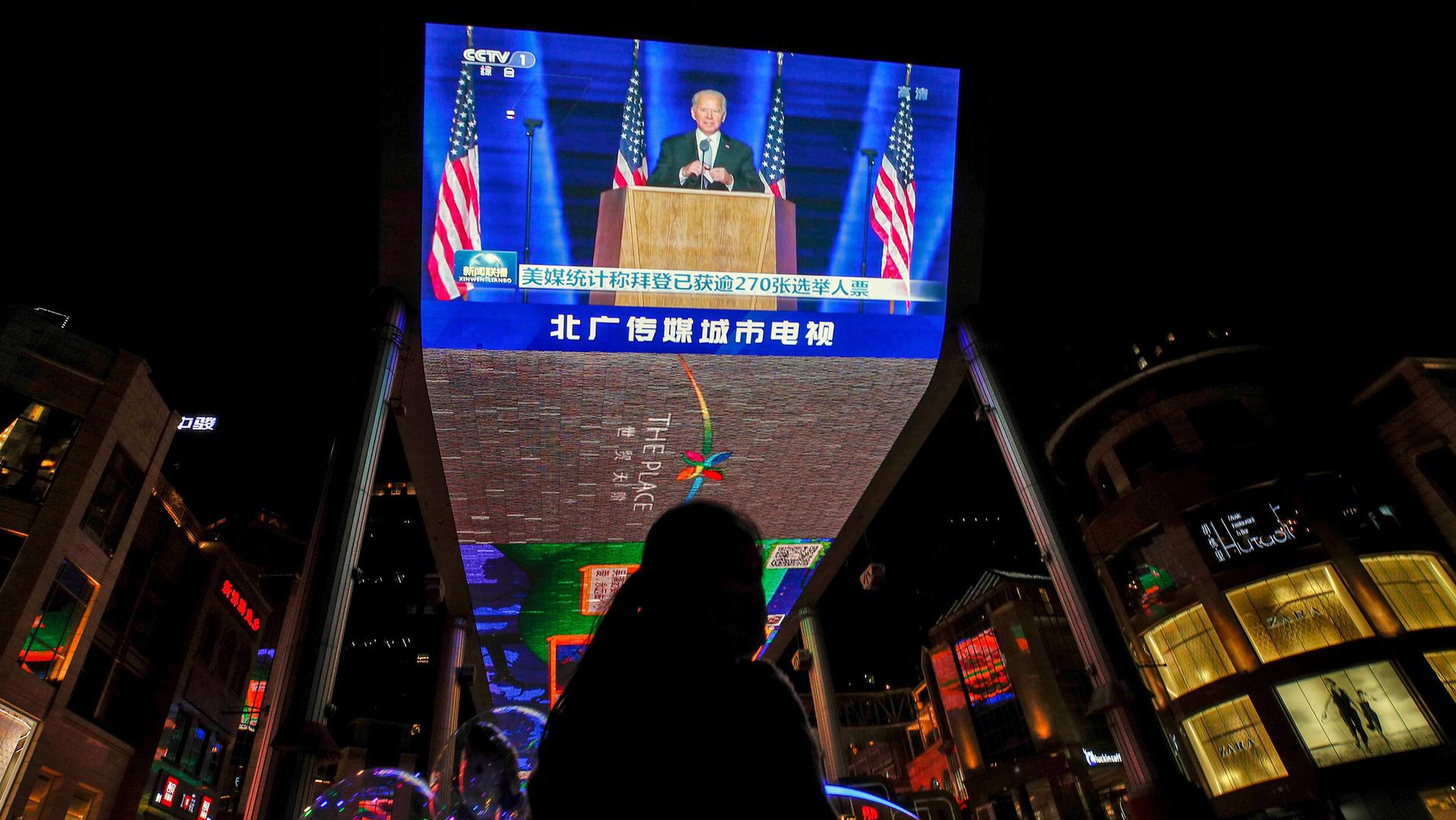 A woman sells balloons near a giant TV screen broadcasting news of US President-elect Joe Biden delivering his speech, at a shopping mall in Beijing, Nov. 8, 2020.