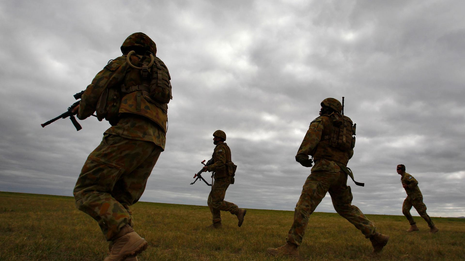 A group of four soldiers are shown walking across a field wearing full battlefield fatigues and carrying weapons.