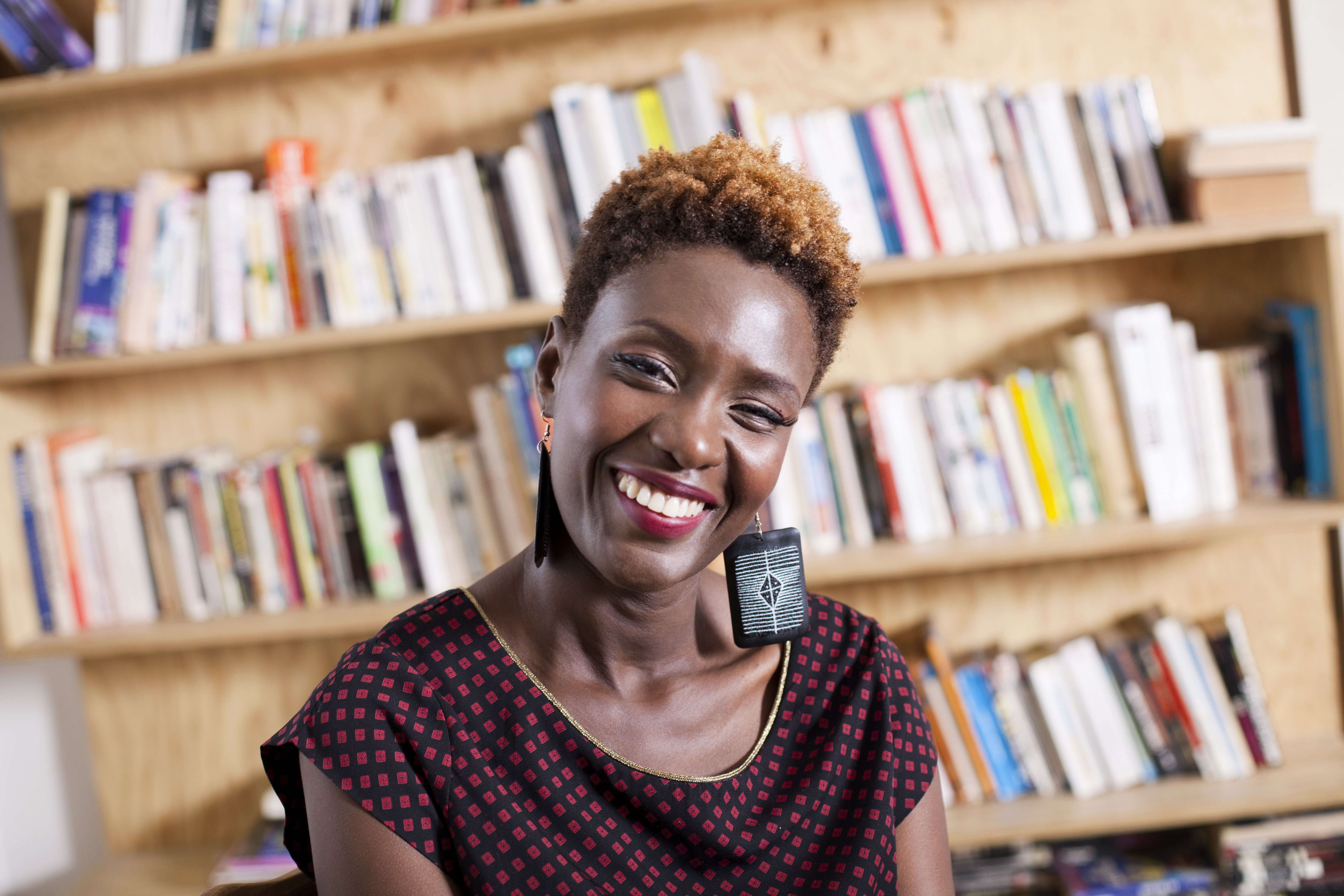A Black woman with short hair wears earrings and a black and red blouse and smiles warmly in front of a large bookshelf.