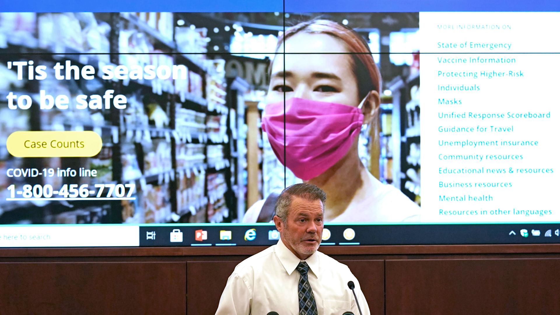 A man wearing a white shirt and gray tie is shown speaking a t podium with a screen behind him showing promotional material on coronavirus safety.