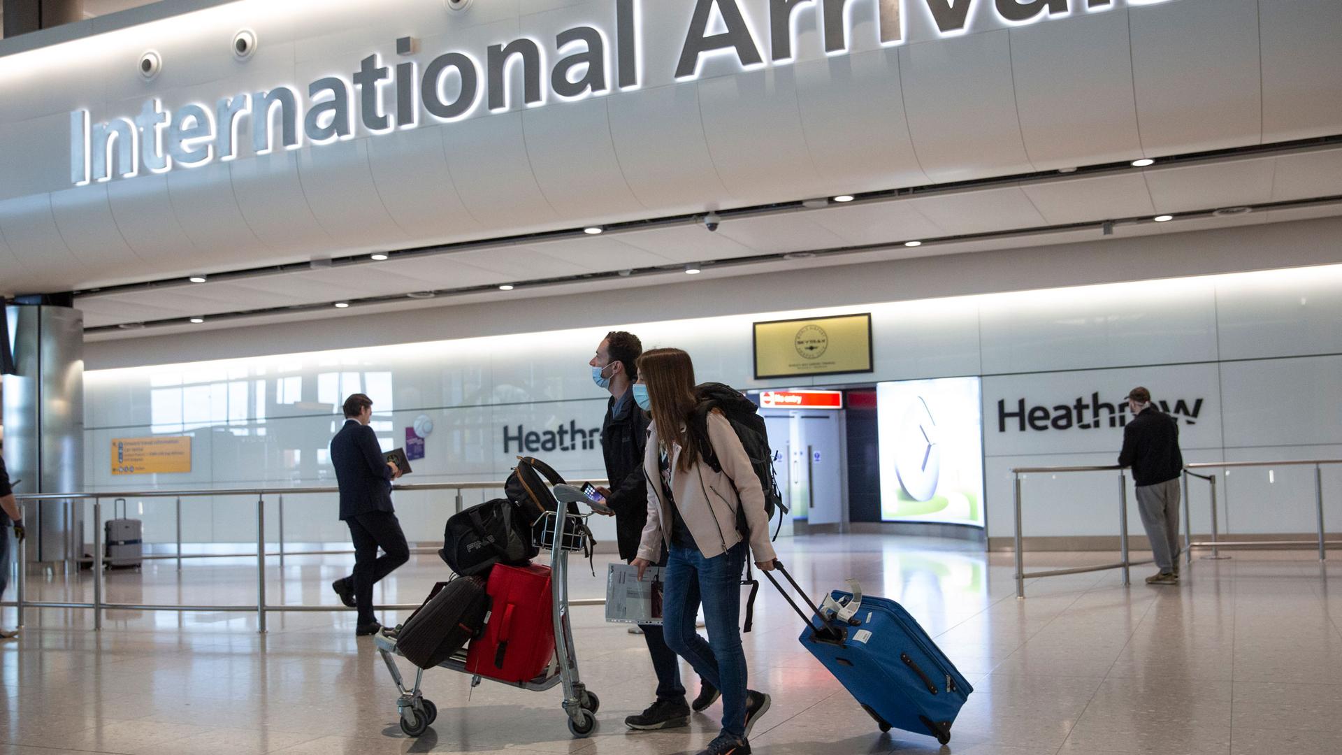 A pair of travelers are shown walking in the International Arrivals section of London's Heathrow Airport with one person pushing a cart carrying several bags.