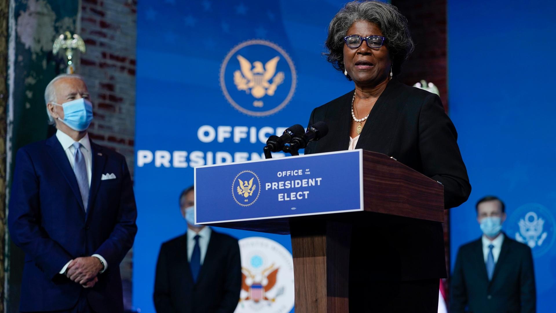 Joe Biden, a white man, is stands near nominee Linda Thomas-Greenfield, a Black woman, who is wearing a dark blue suit onstage at a podium. 