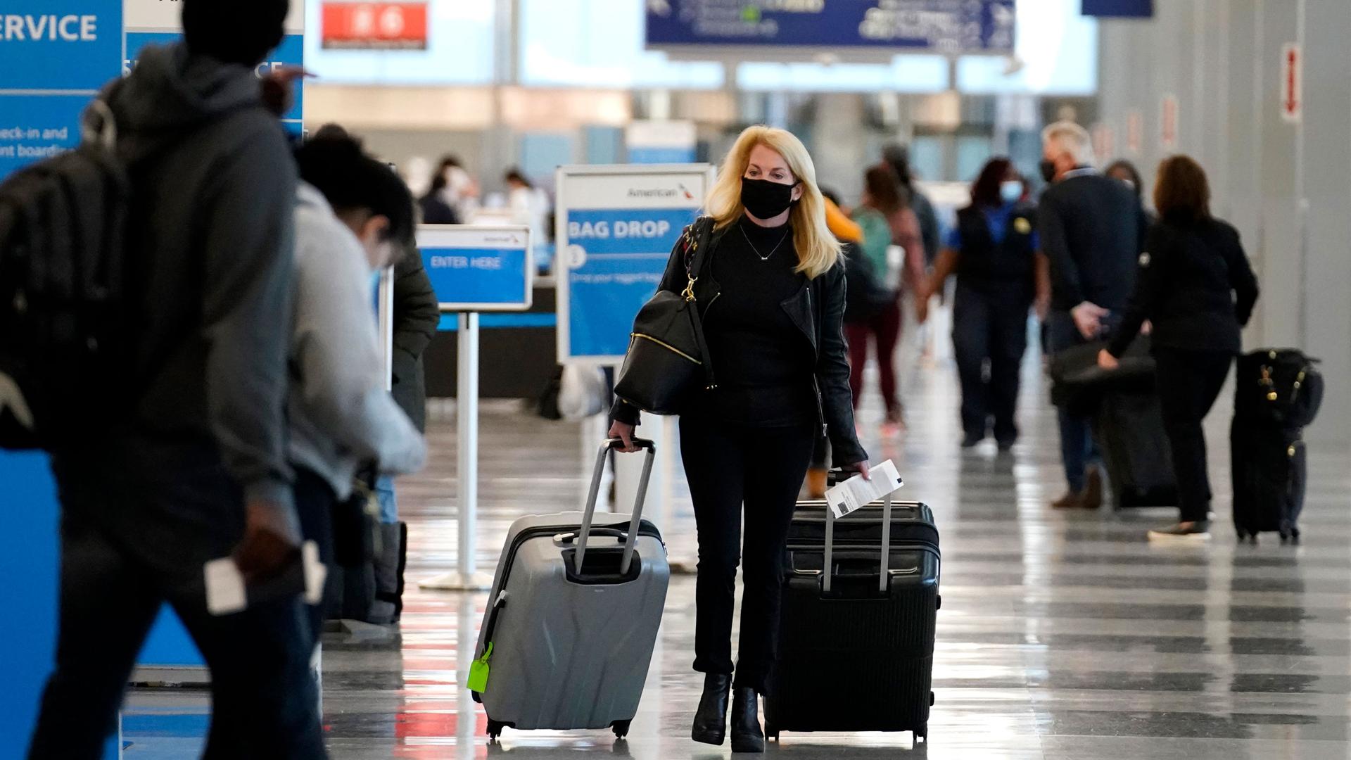 A woman is shown center frame pulling two rollaboard suitcases while wearing a face mask.