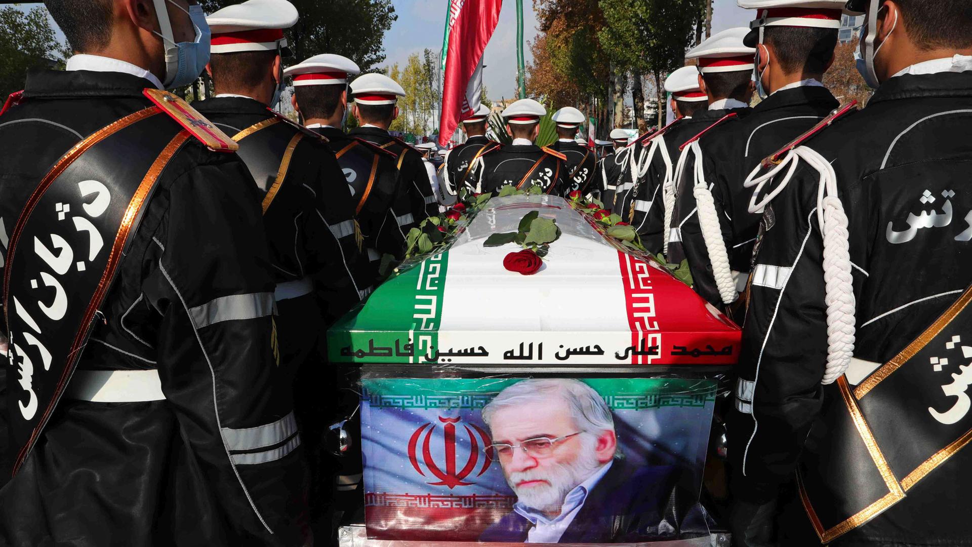 Rows of military personnel are shown on either side of a coffin that has an Iranian flag on top and a photo of Mohsen Fakhrizadeh on the side.